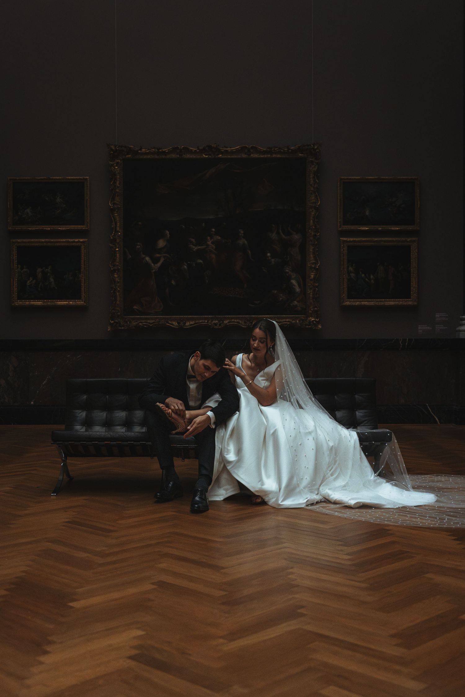 Wedding couple seated in art gallery with dramatic lighting and herringbone floor.
