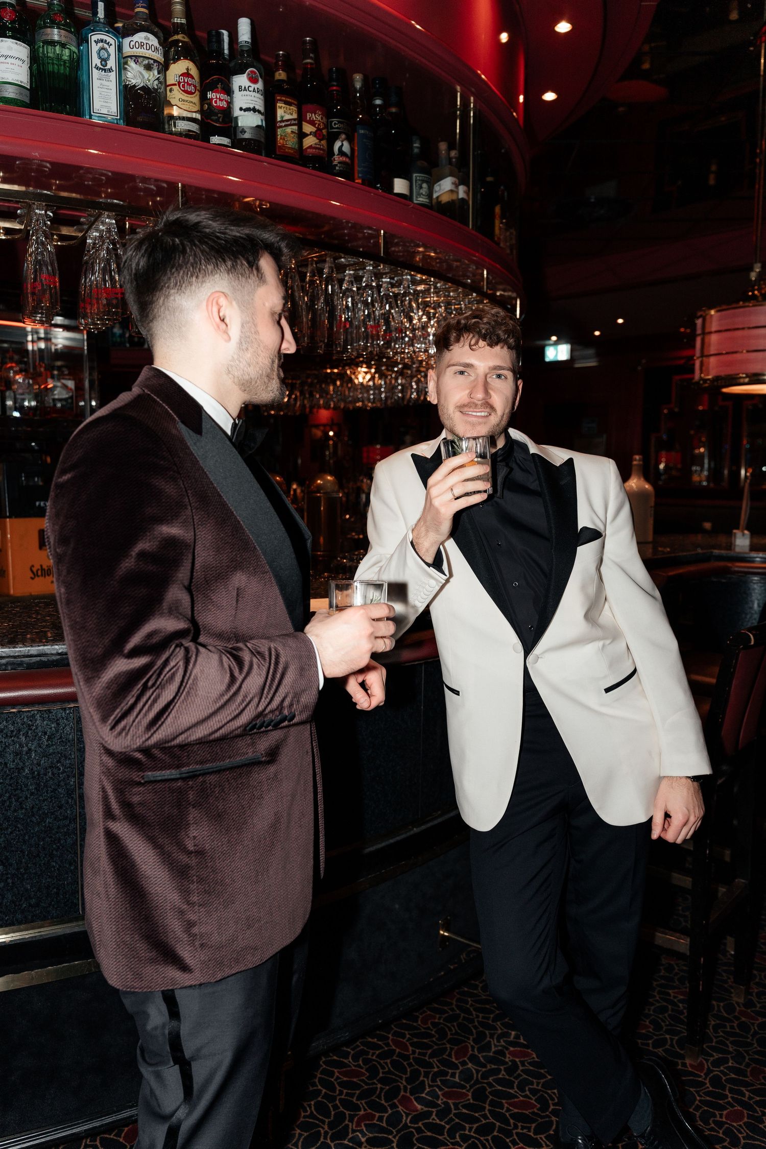 Two men in formal attire having drinks at a stylish bar with red lighting and bottles on display.