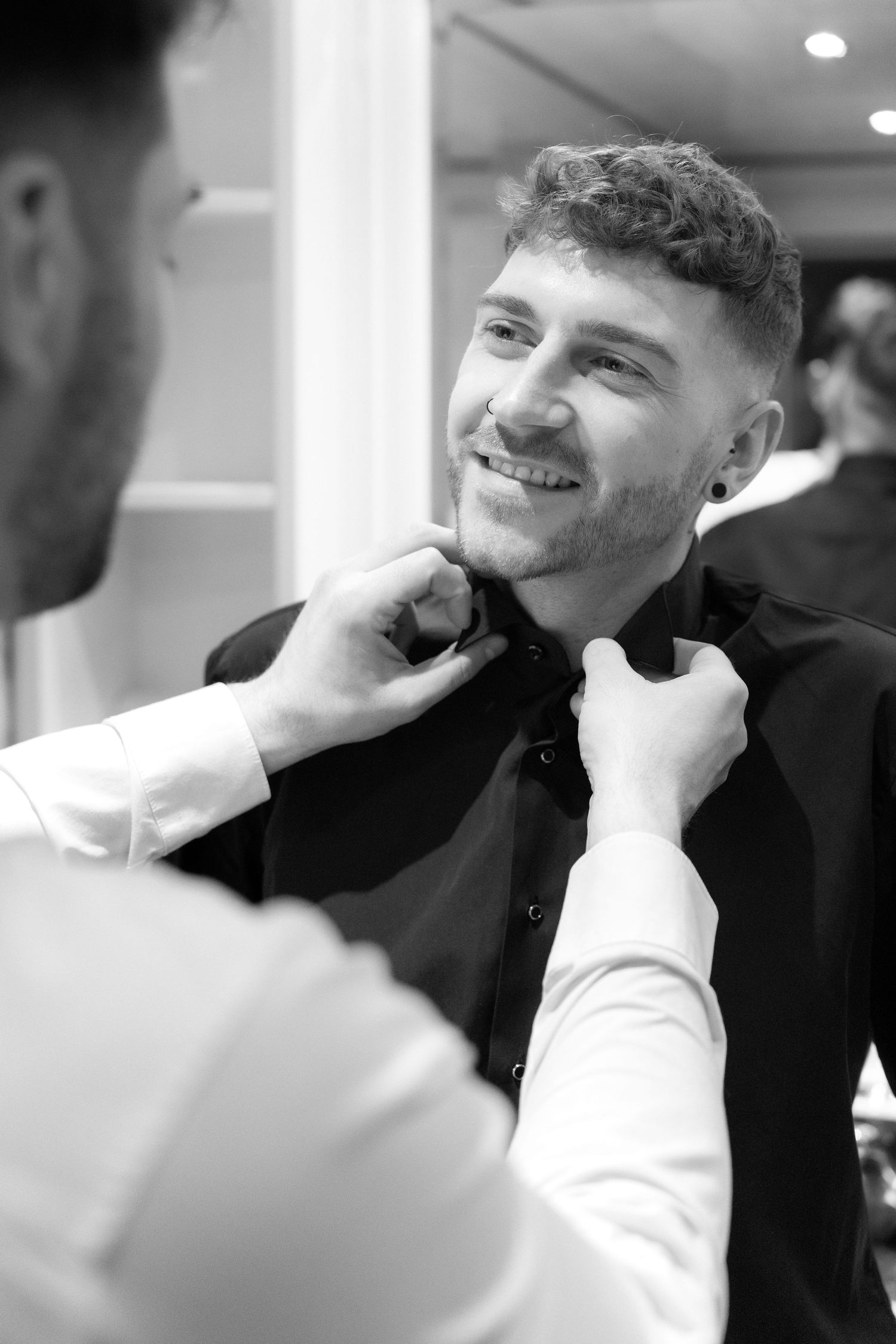 Black and white photograph showing someone having their bowtie adjusted while smiling warmly in formal attire.