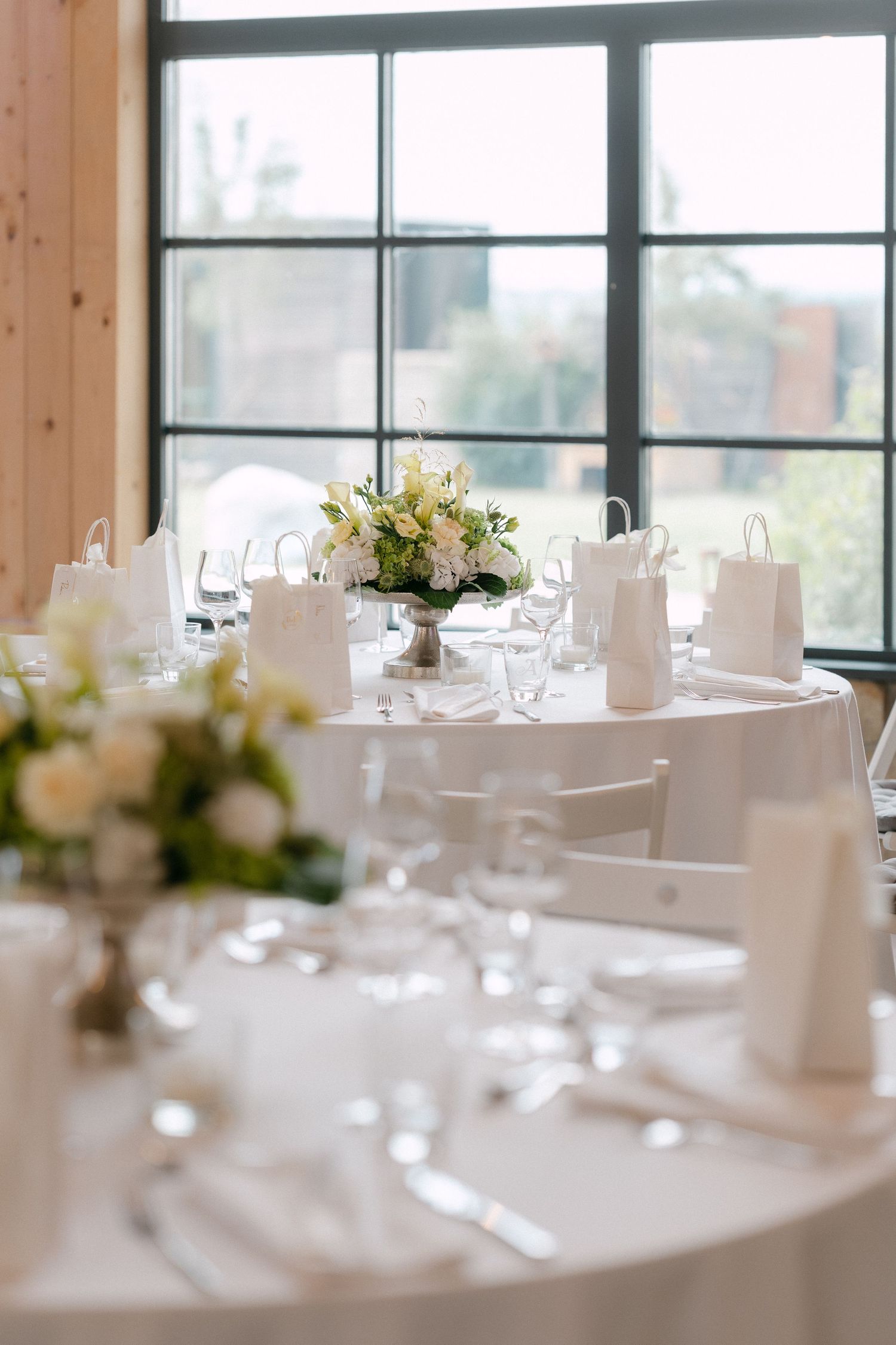 Elegant wedding reception tables with white linens and floral centerpieces set against large windows in a bright venue.