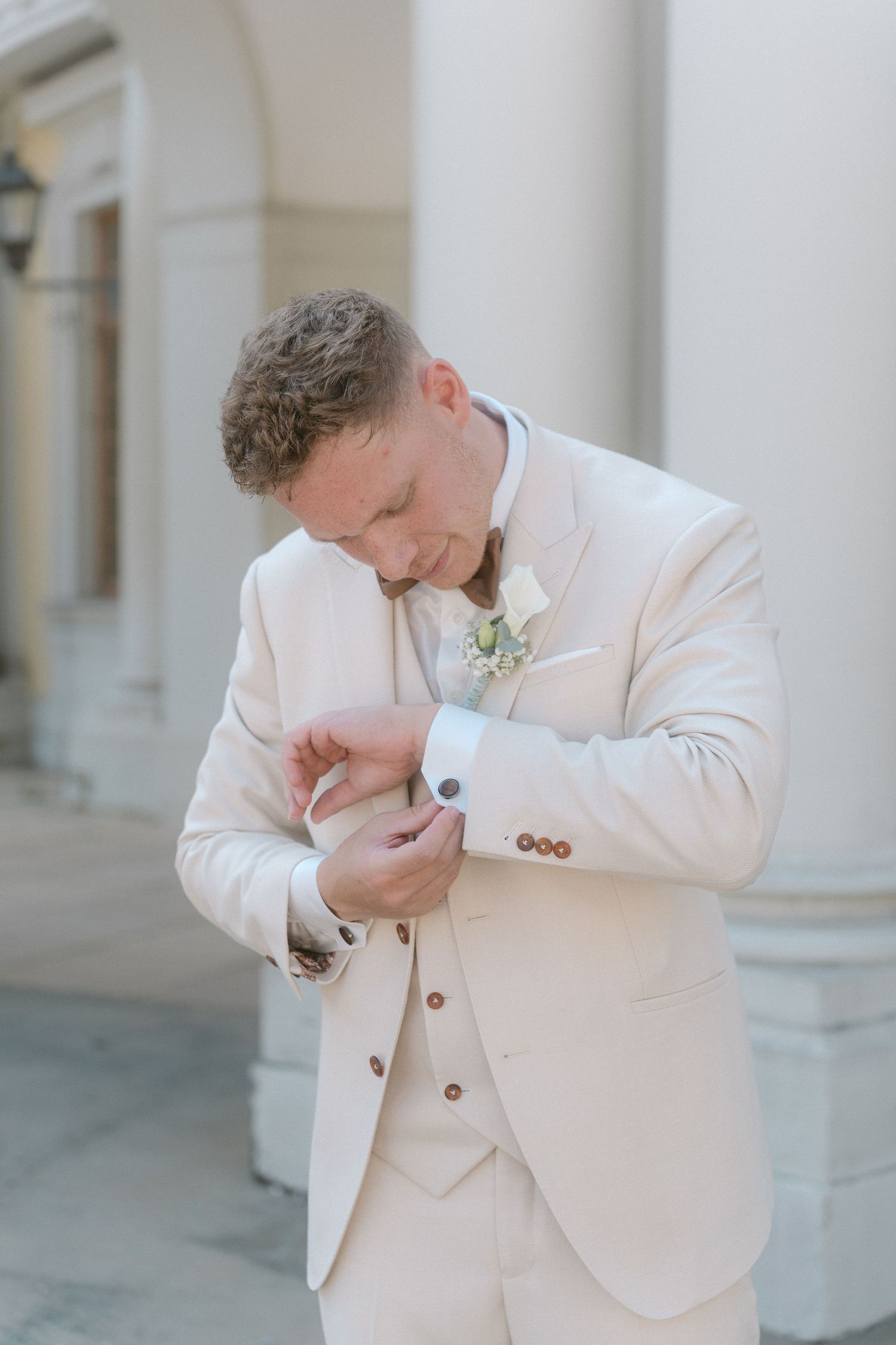 A person in a white suit adjusts their boutonniere while standing in front of classical columns. Schloss Ludwigsburg