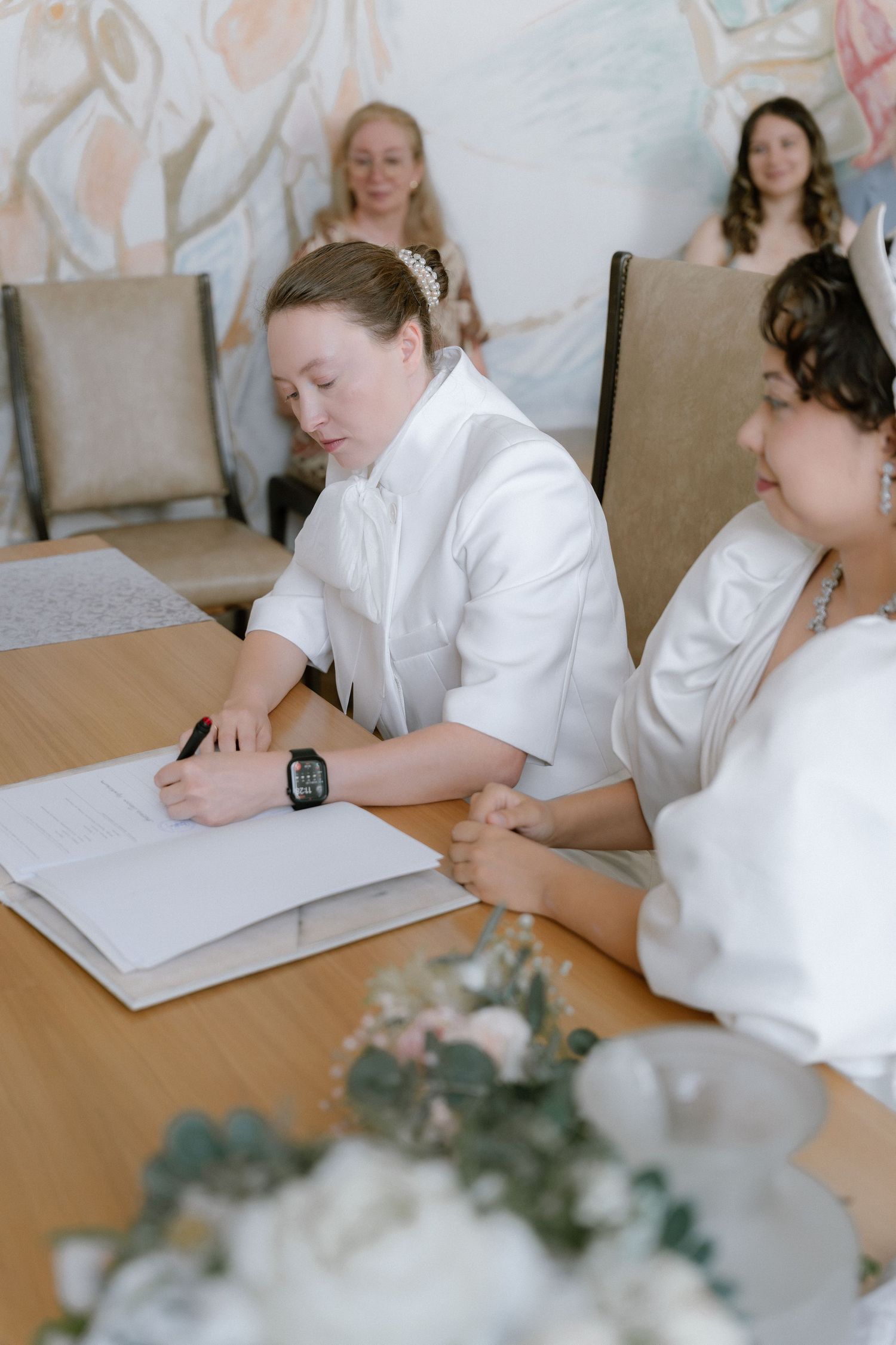 Two brides in white signing wedding registry documents at a table during civil ceremony.