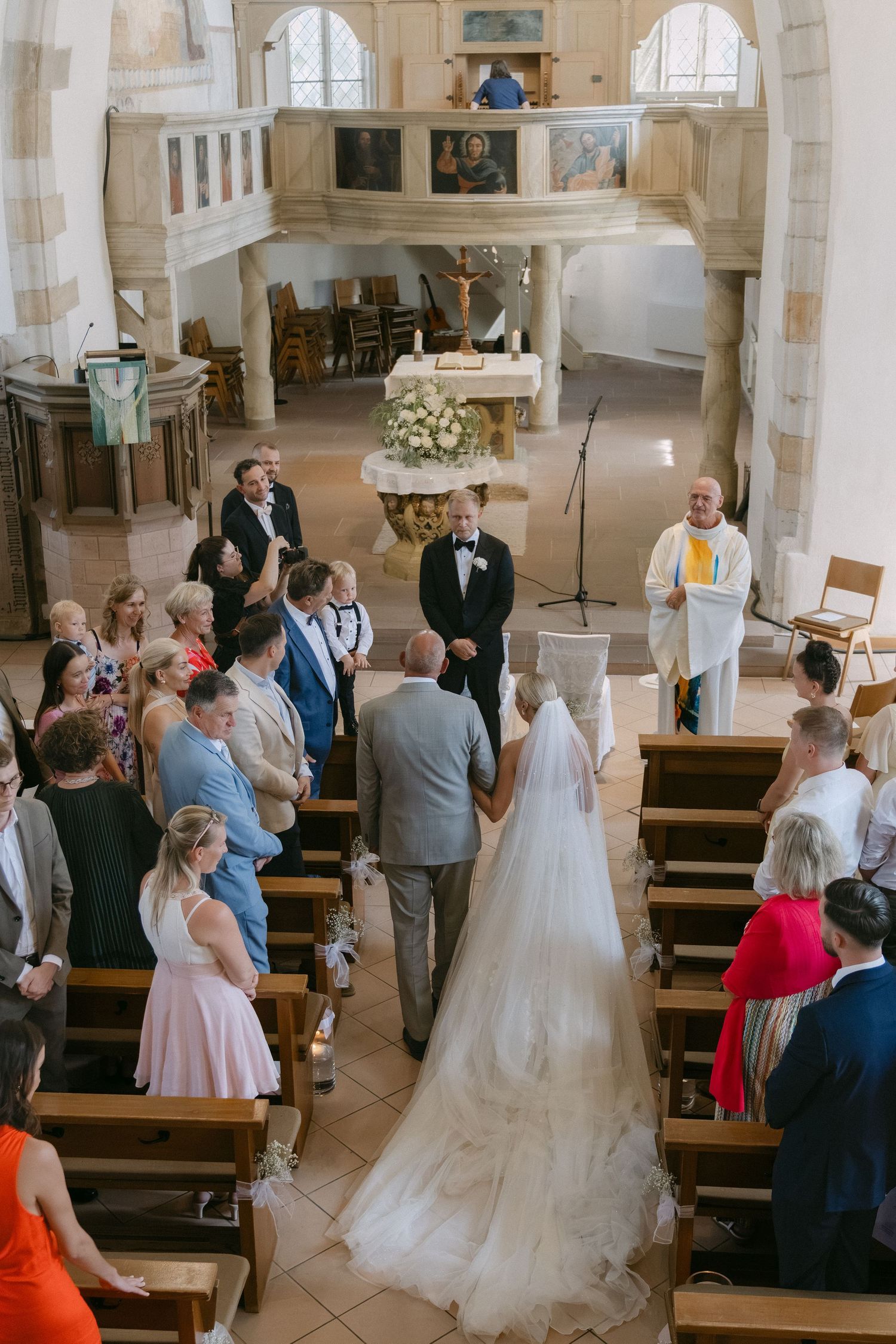 Bride in a long veil walks down the church aisle toward the groom and officiant as seated wedding guests look on.