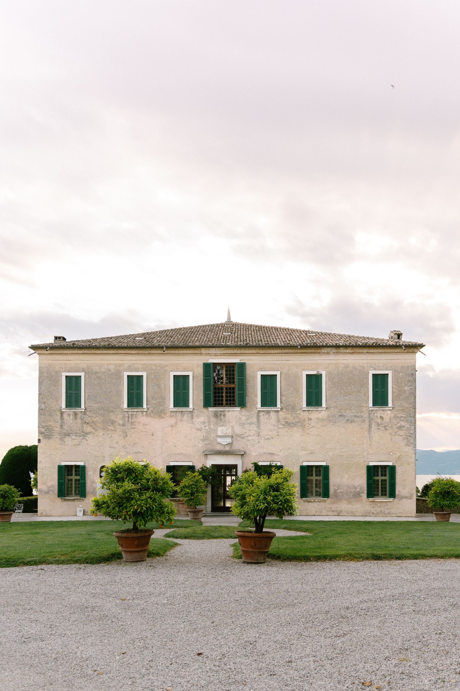 Rustic Mediterranean villa with green shutters and potted plants on a gravel courtyard against cloudy skies.