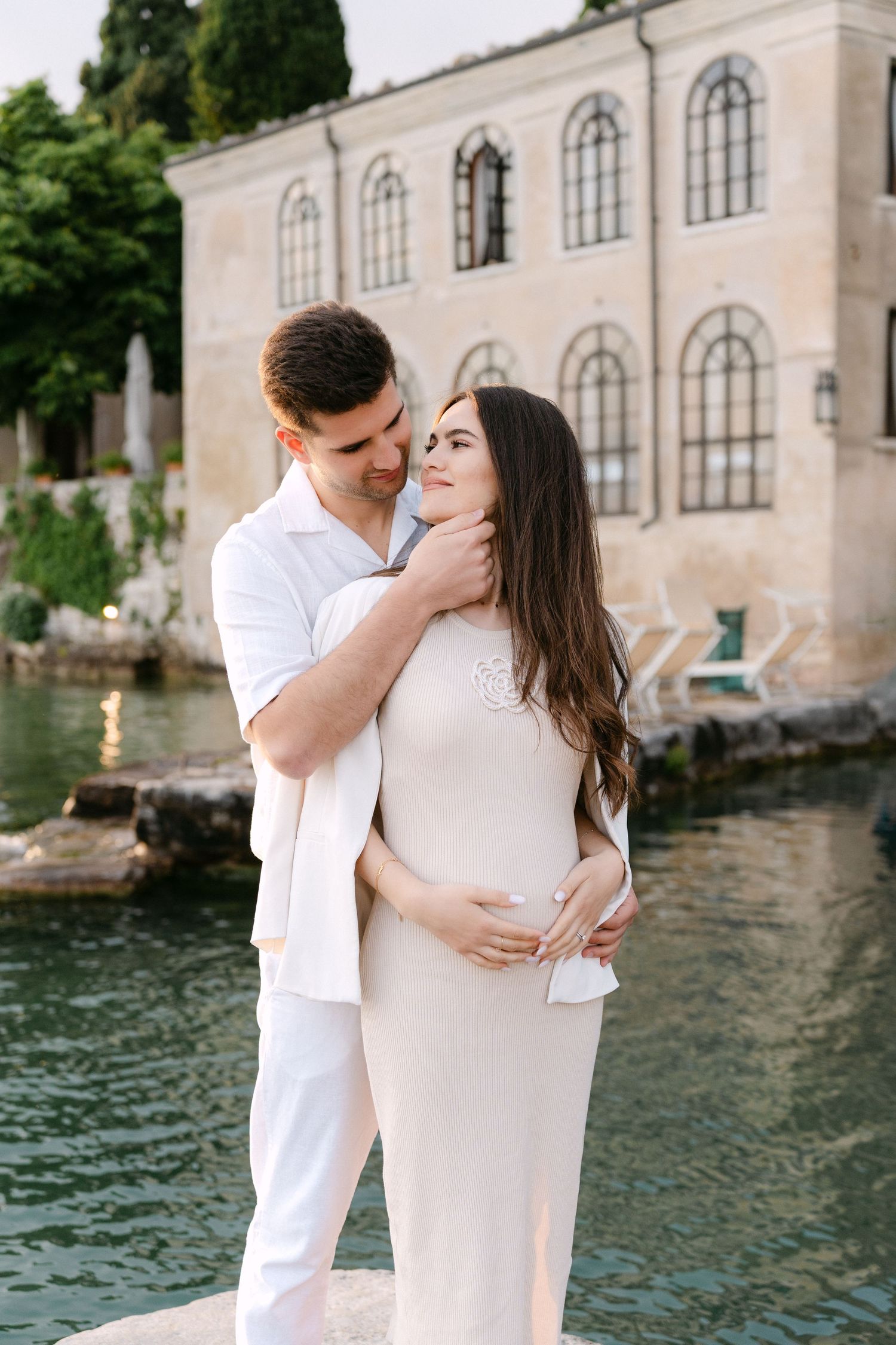 A couple in white outfits embraces by the water near a historic building with arched windows in Italy.