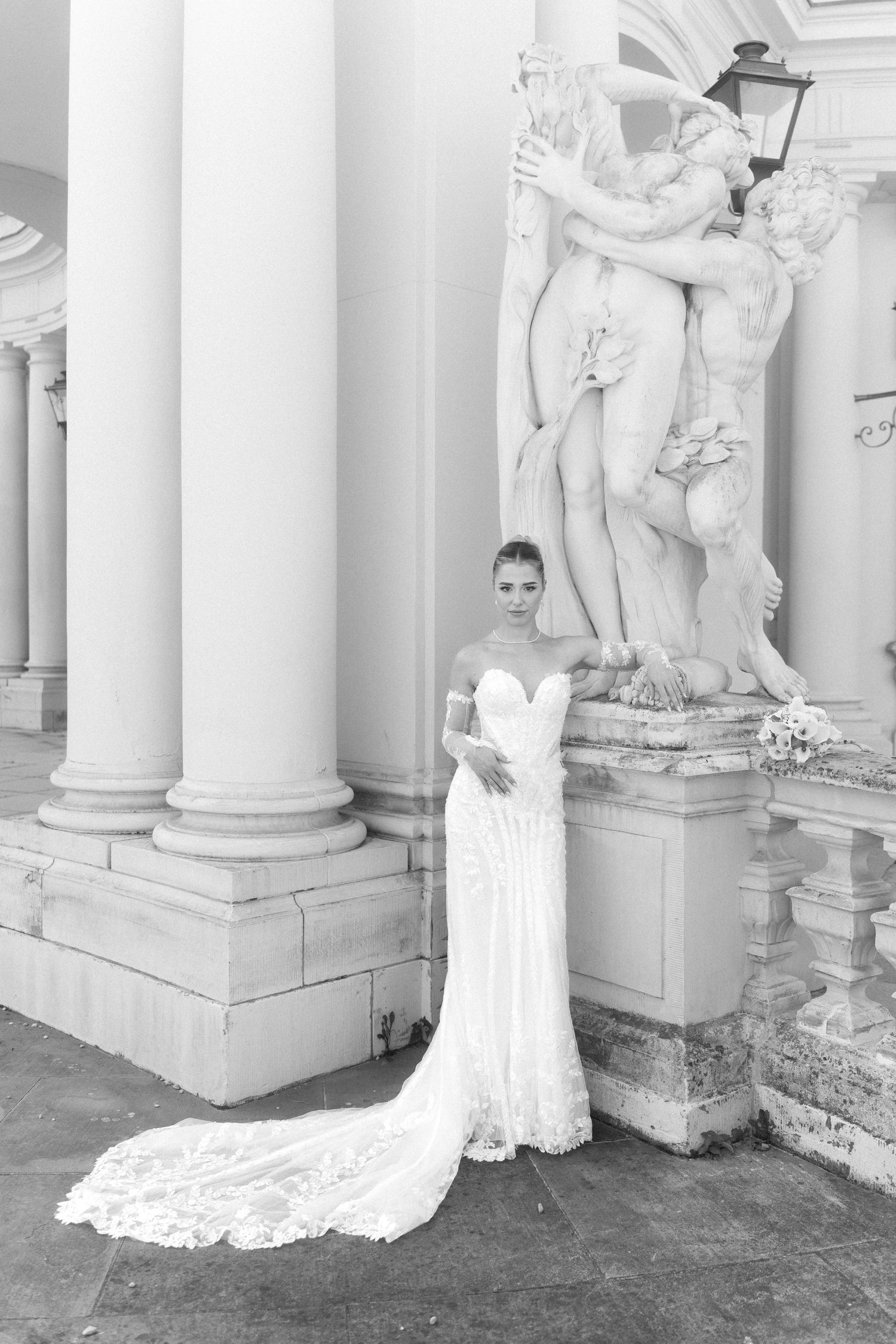 Elegant white wedding dress with long lace train photographed in front of classical marble statues and columns. Schloss Ludwigsburg