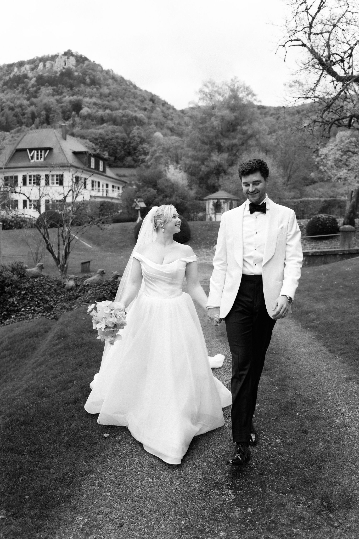 A couple in wedding attire walks together along a scenic countryside path in black and white photography.