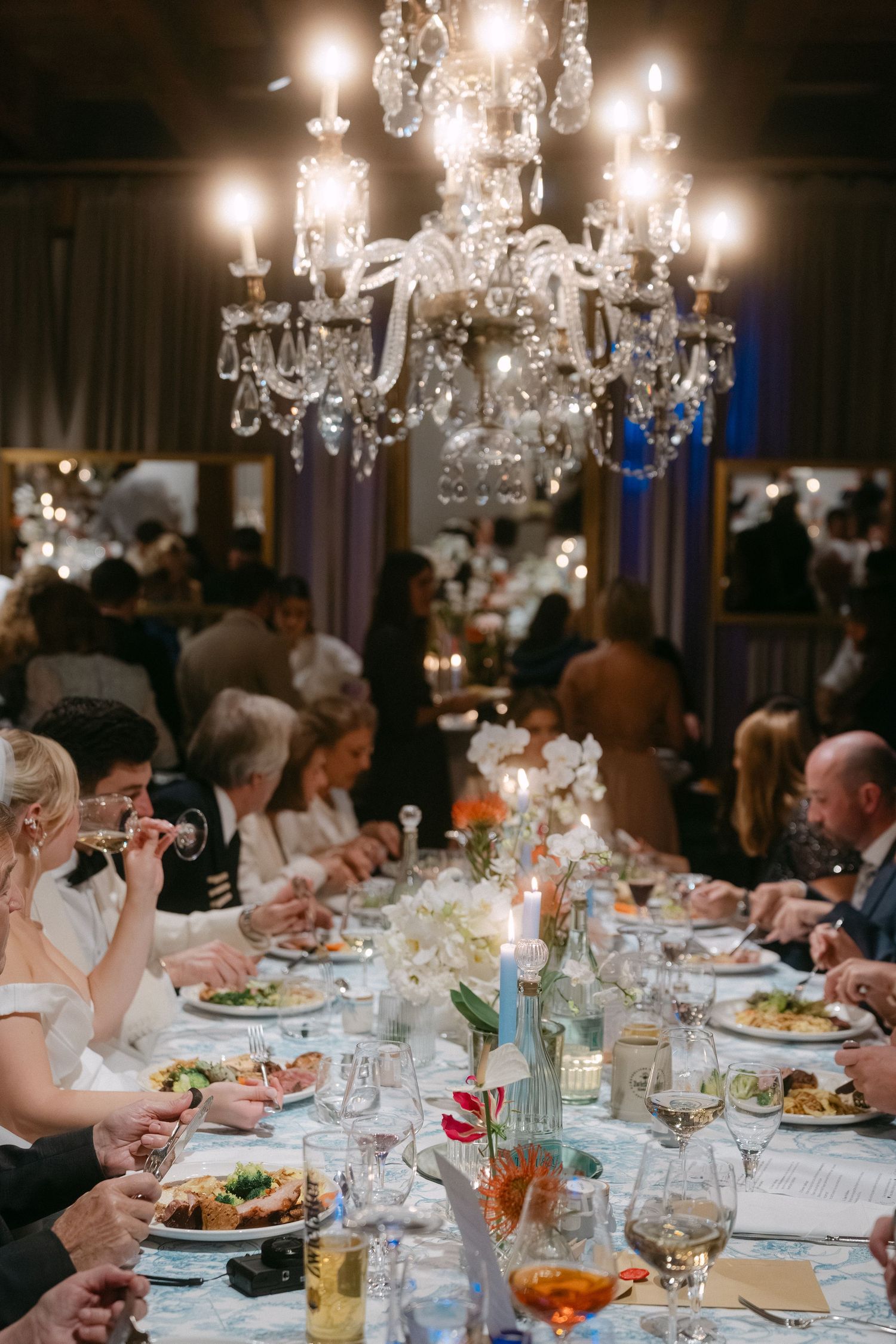 Elegant crystal chandelier illuminates formal dinner reception with guests seated at long tables decorated with candles and flowers.