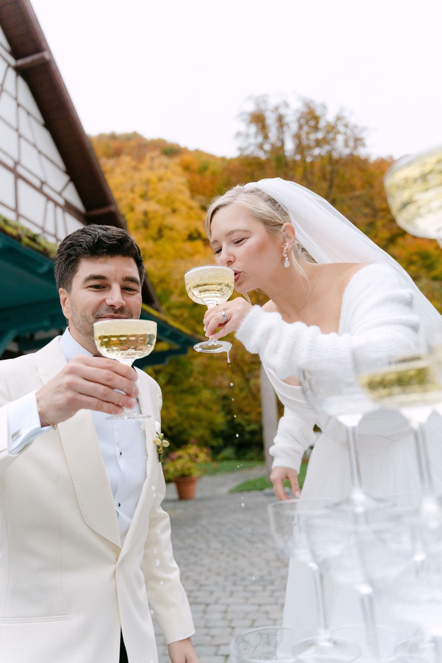 A bride and groom in white attire toast with champagne glasses against an autumn backdrop of golden trees.