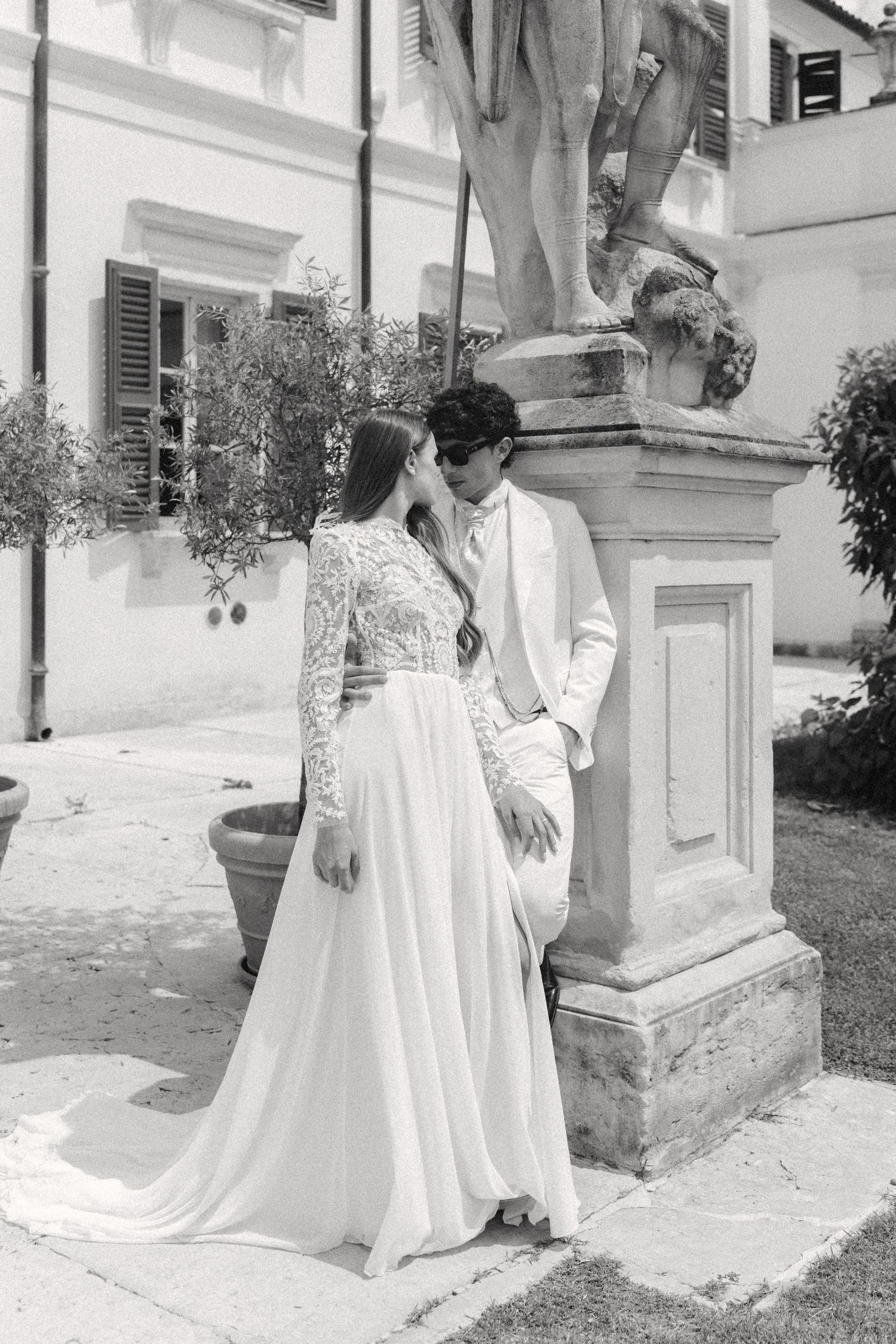 A romantic black and white wedding photo of a couple in elegant attire posing by a classical stone pillar in a garden setting. Villa Mosconi