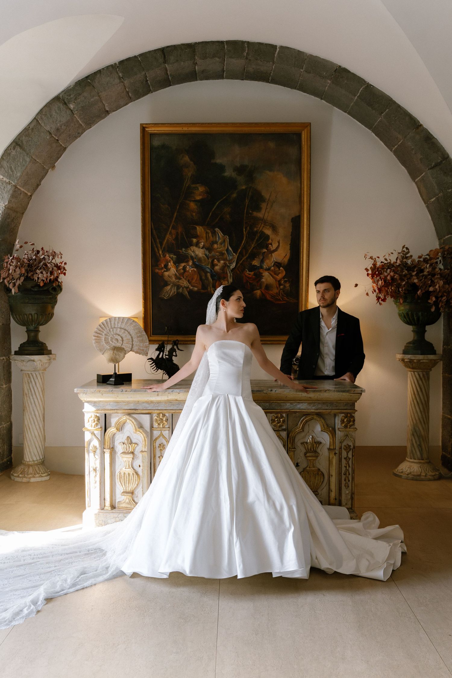 Bride in white ballgown stands under stone arch with groom behind ornate altar and classic painting in elegant venue.
