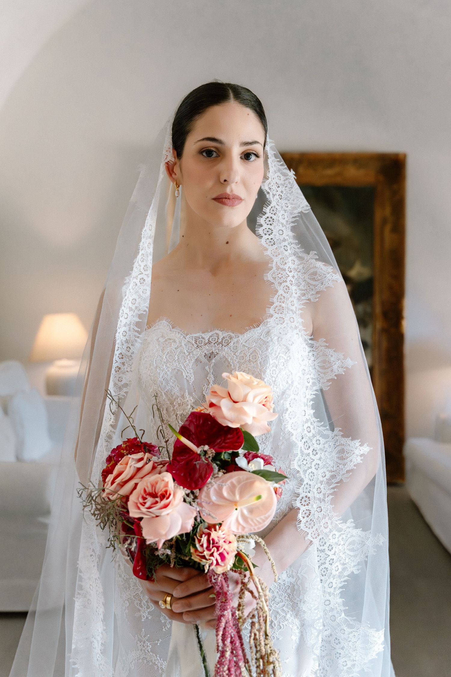 Bride in lace wedding dress and veil holds pink and red rose bouquet indoors.