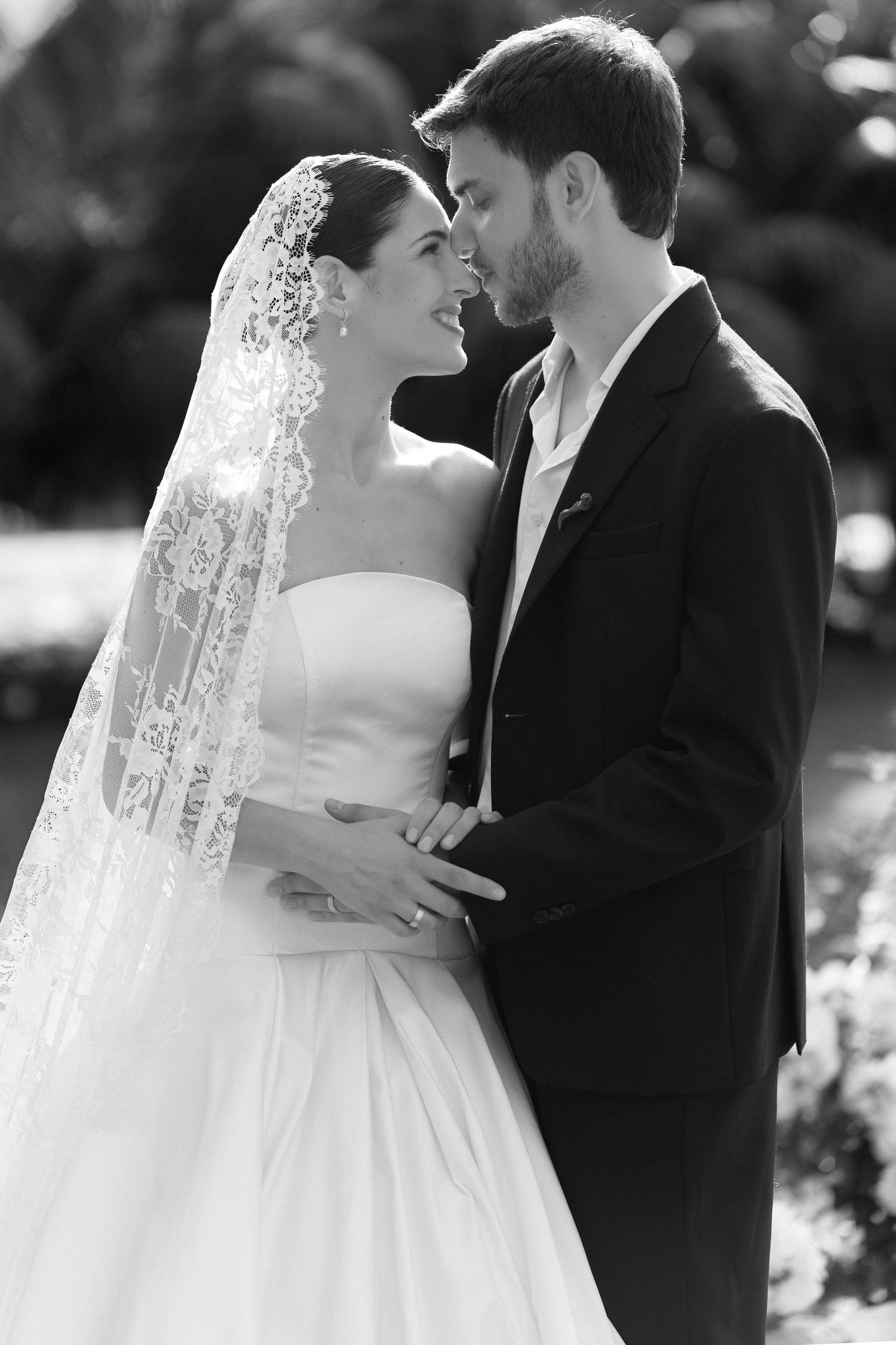 Black and white photo of a bride in lace veil and groom in suit sharing a romantic moment outdoors.