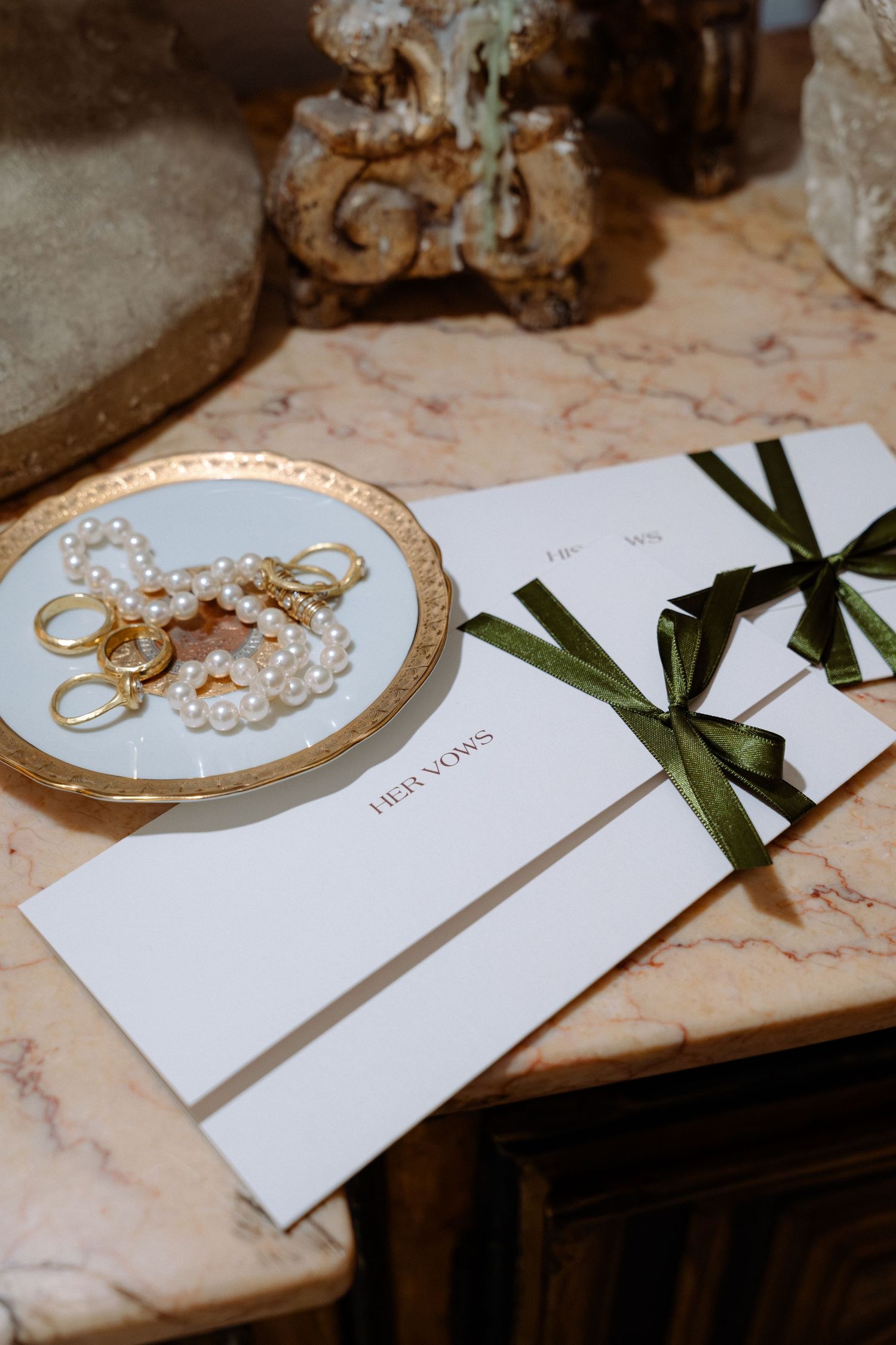 Elegant wedding stationery with green ribbon bows beside a gold plate with pearl jewelry and rings on marble.