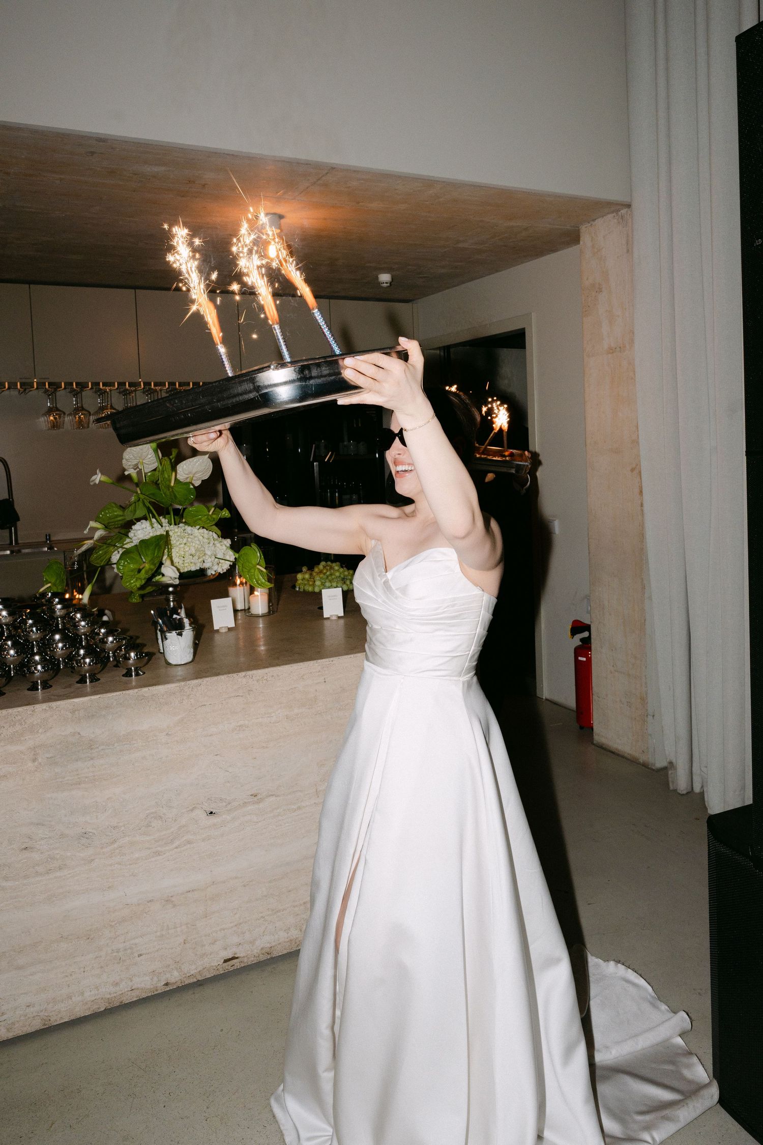 Bride in white strapless gown holds sparkling cake above her head at elegant wedding reception venue.