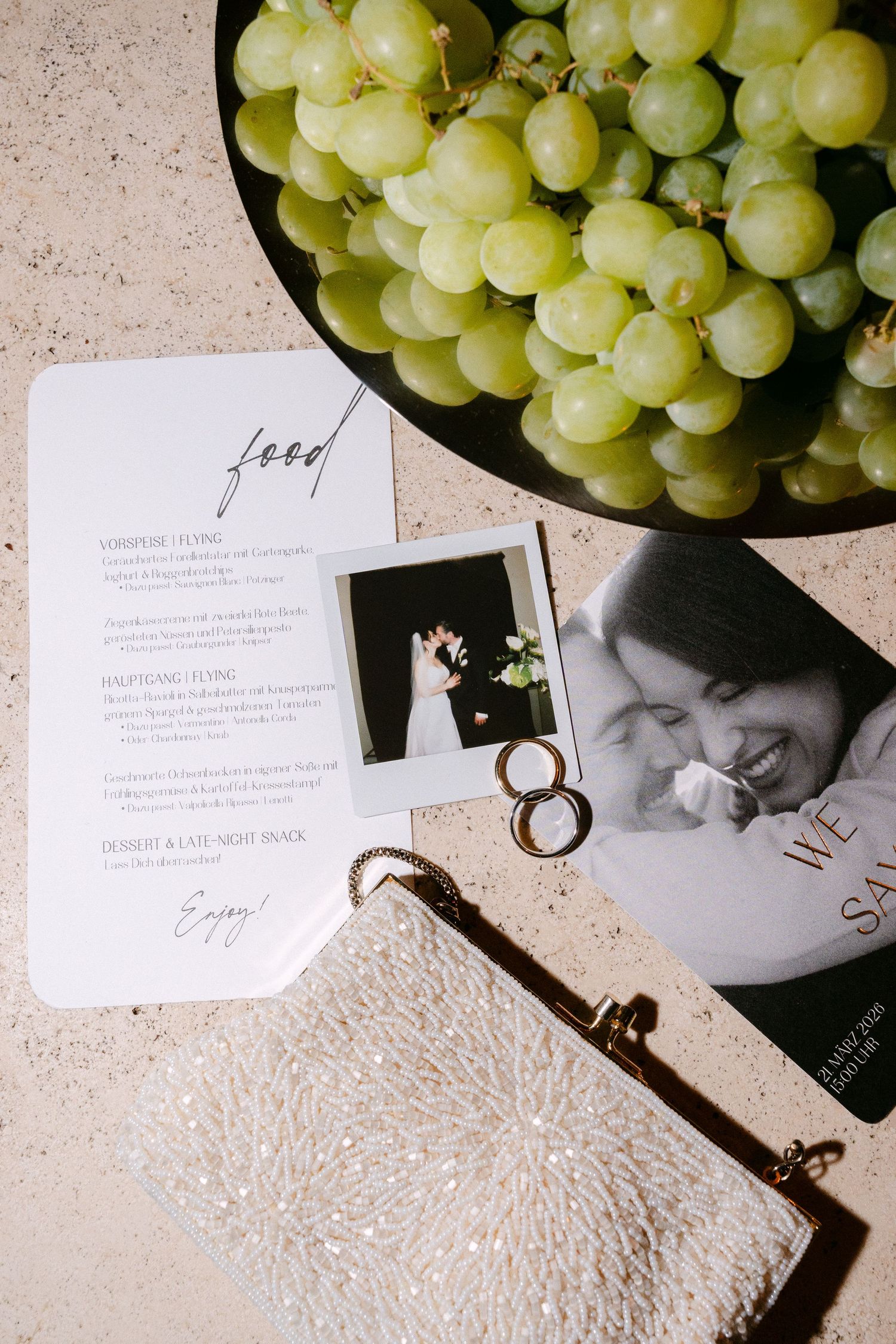 Wedding flat lay with food menu, Polaroid photo, gold rings, beaded clutch, and fresh green grapes on a plate.