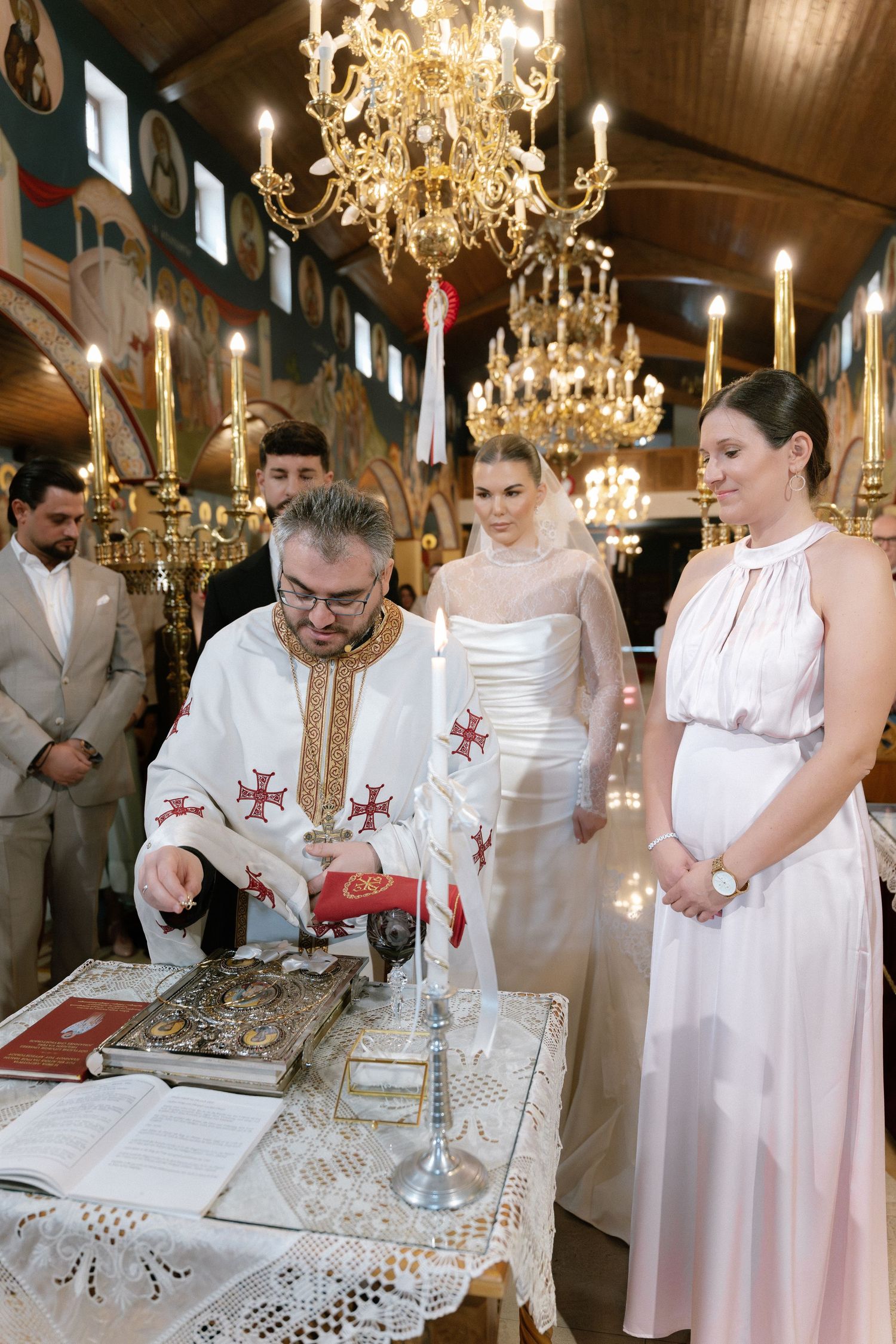 Orthodox priest performs wedding ceremony with bride and bridesmaid at ornate church altar with chandeliers.