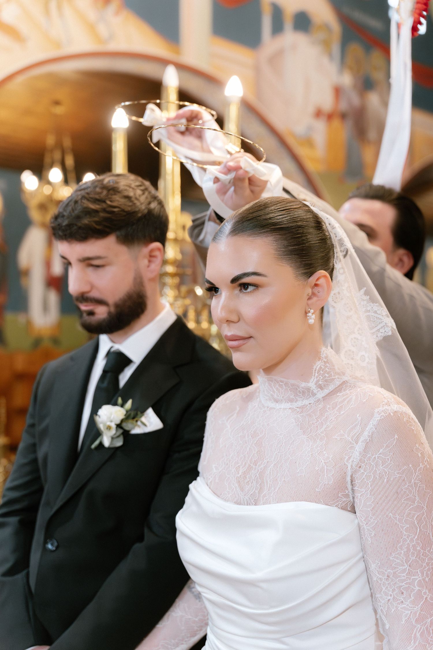 Bride in white lace gown and groom in black suit during Orthodox wedding ceremony with crowning ritual.