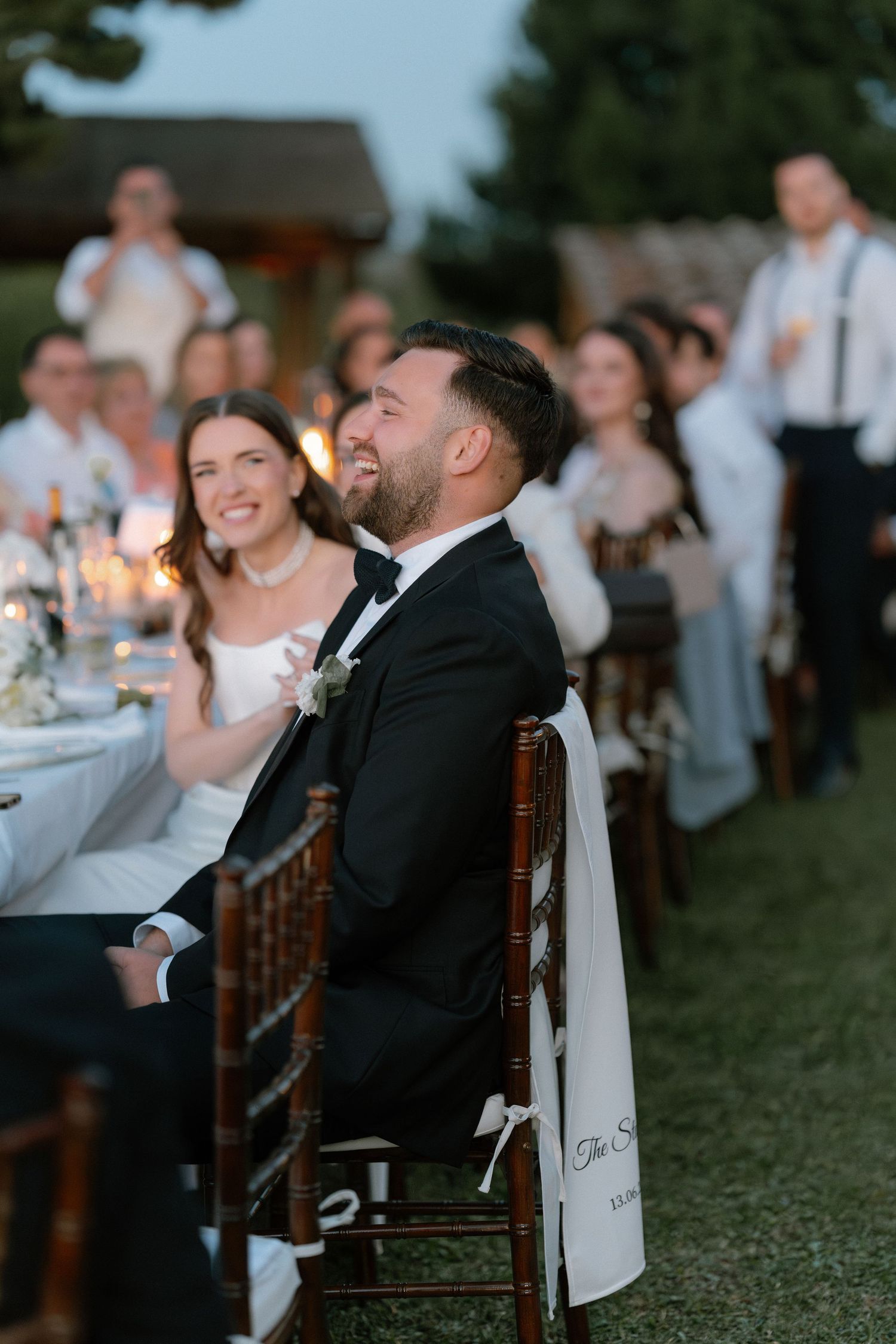 Bride and groom laughing joyfully at outdoor wedding reception dinner, guests seated along long table at dusk.