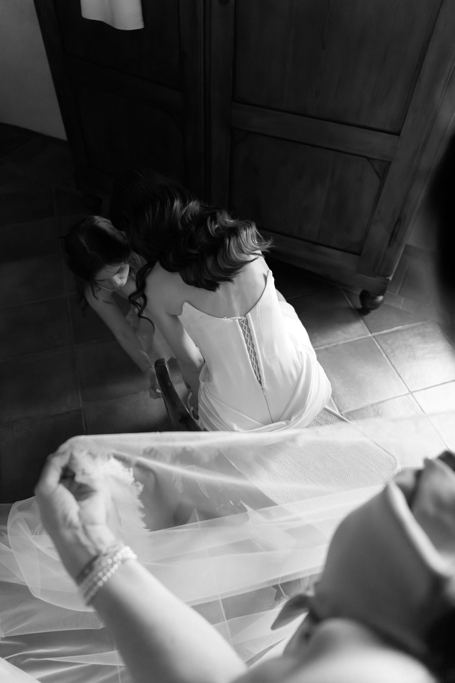 Black and white photo of bride being helped into wedding dress by bridesmaids in a rustic room.