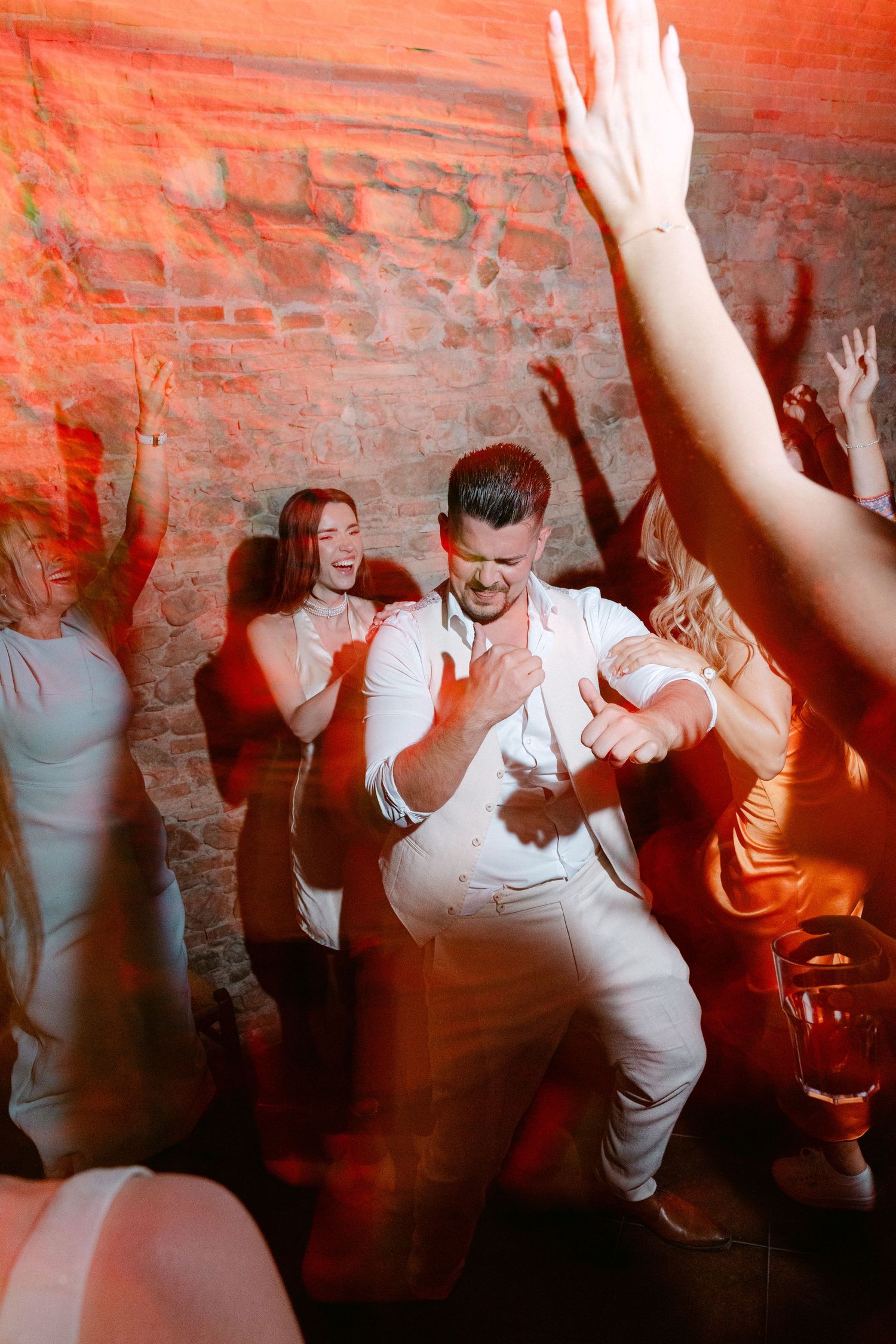 Man in white outfit dancing energetically at a party with friends cheering under red lights near a brick wall.