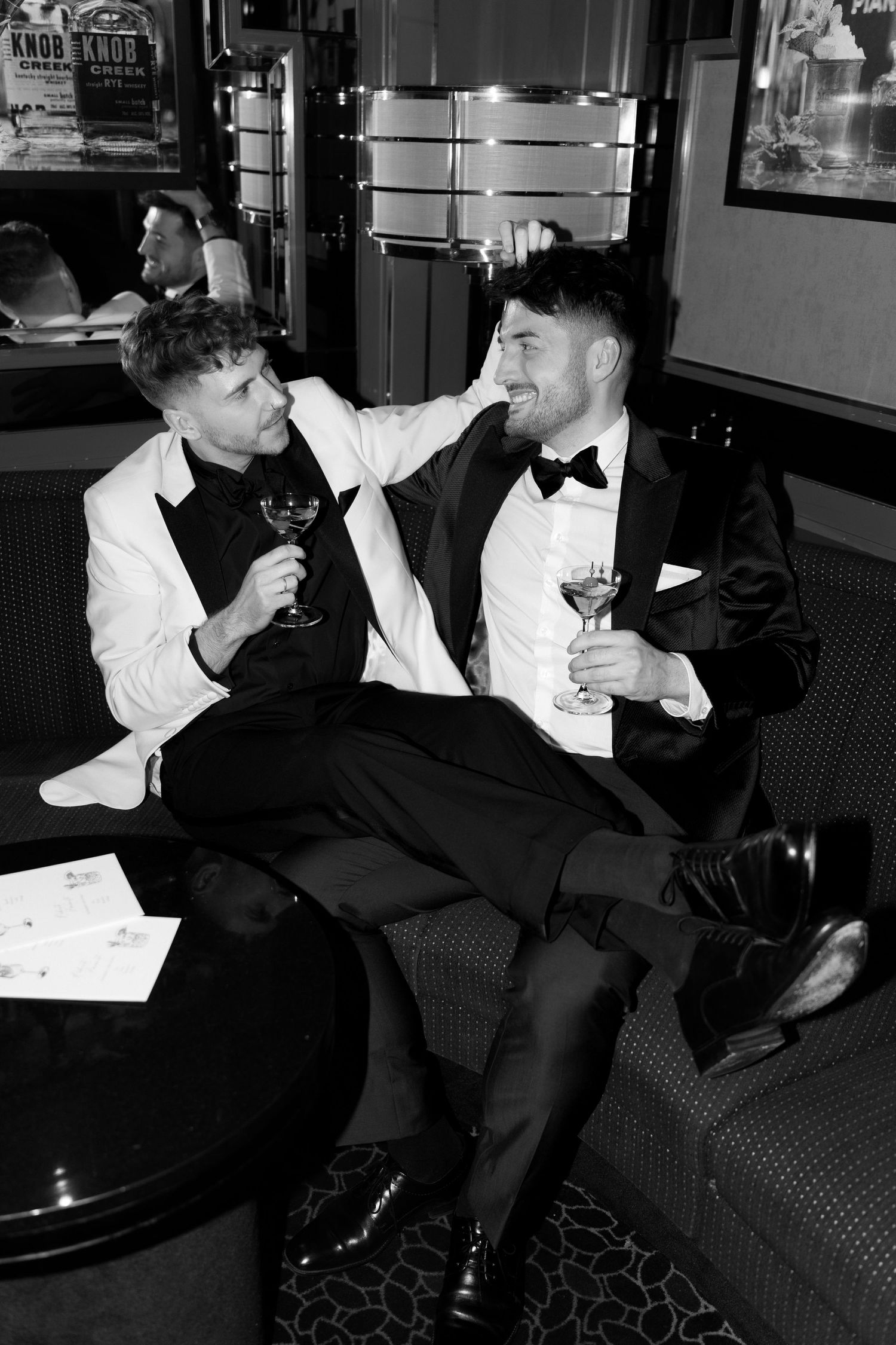 Two people in formal attire with bow ties relax on a couch while enjoying wine at an elegant venue.