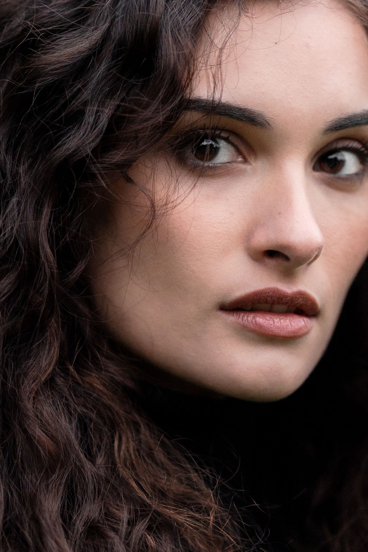 A close-up portrait of a woman with dark wavy hair, red lips and brown eyes looking to the side against a black background.