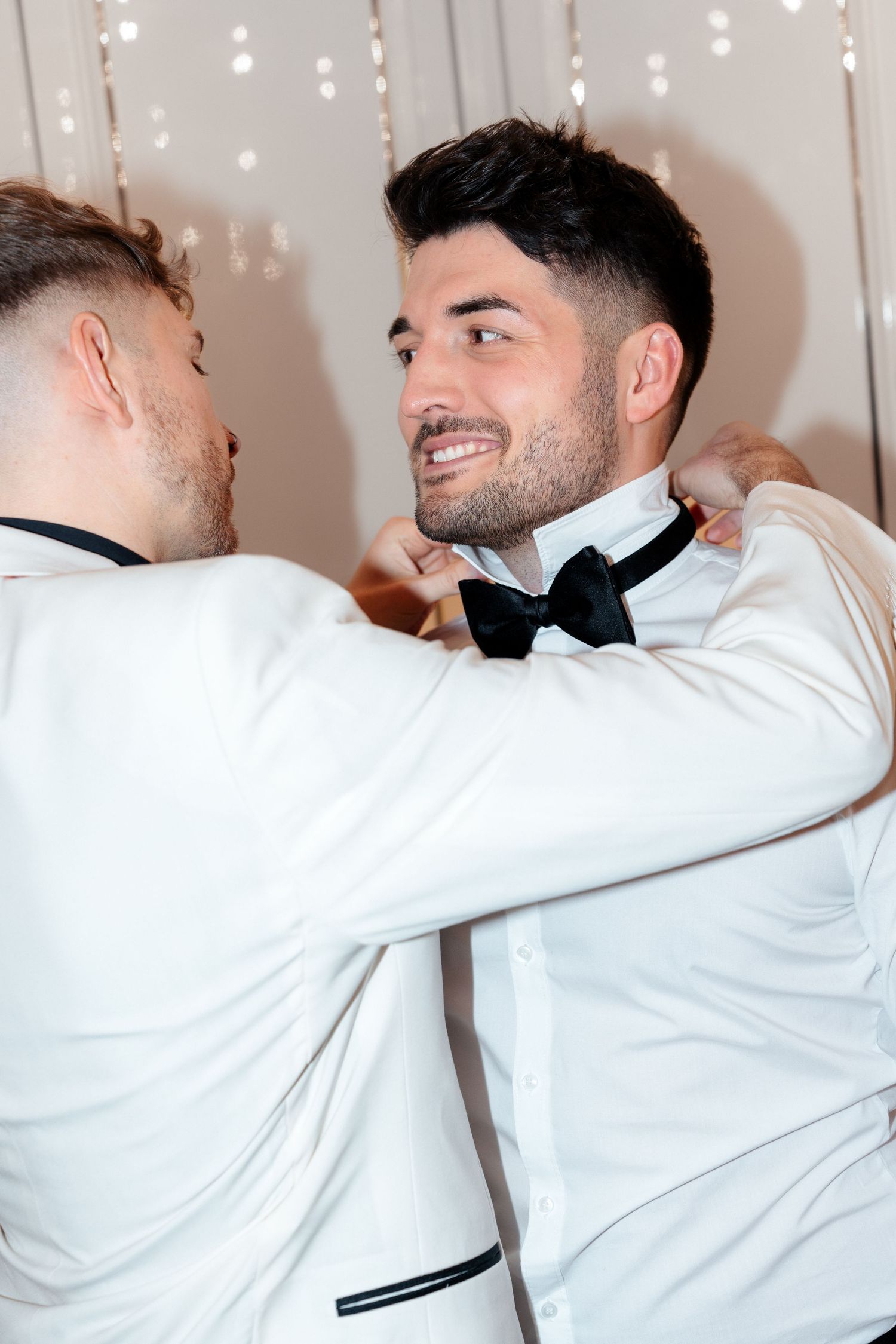 Two people in matching white tuxedos and black bowties embrace while smiling at each other against a sparkly backdrop.