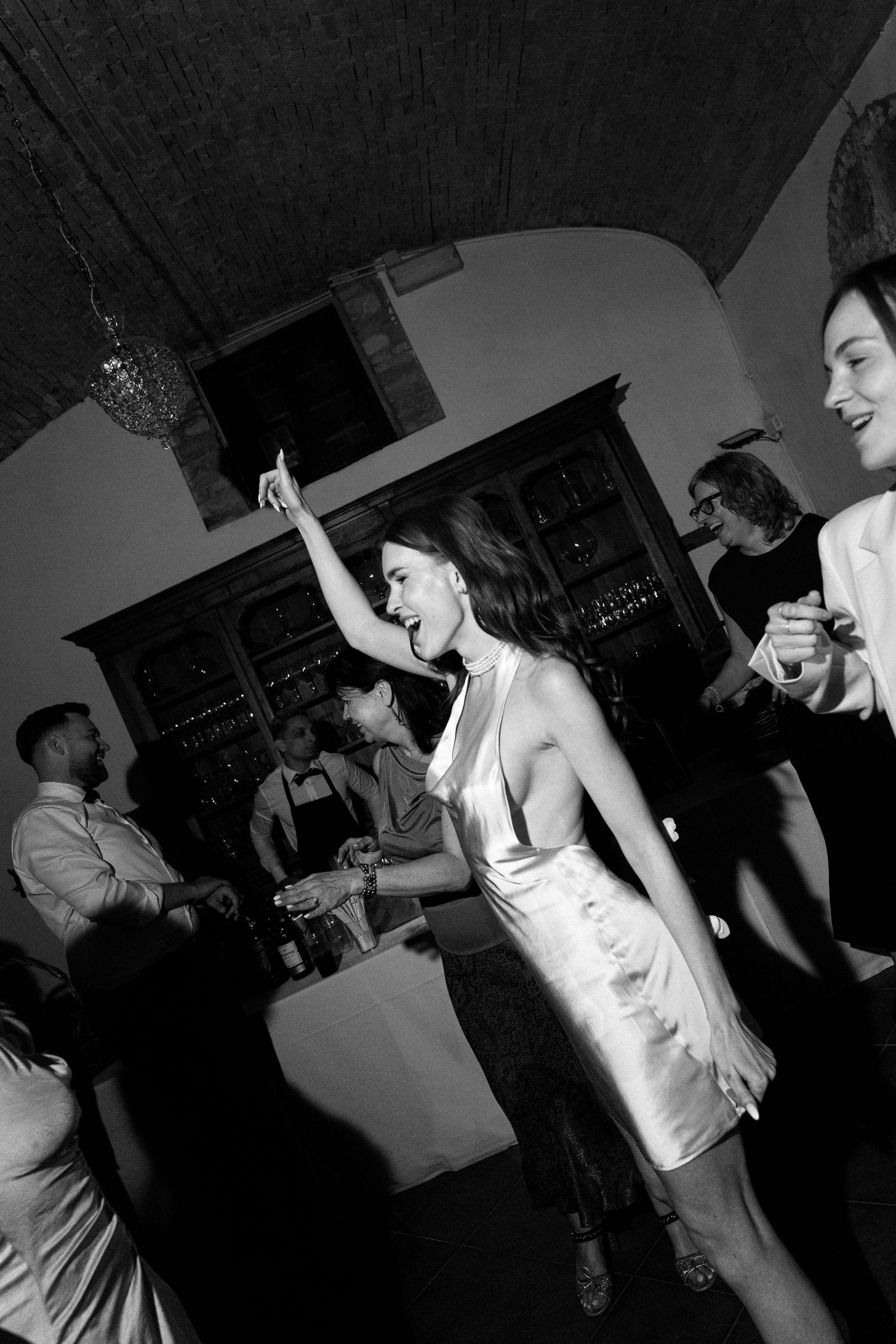 Black and white photo of a woman in a white dress dancing joyfully at an elegant party with guests in the background.