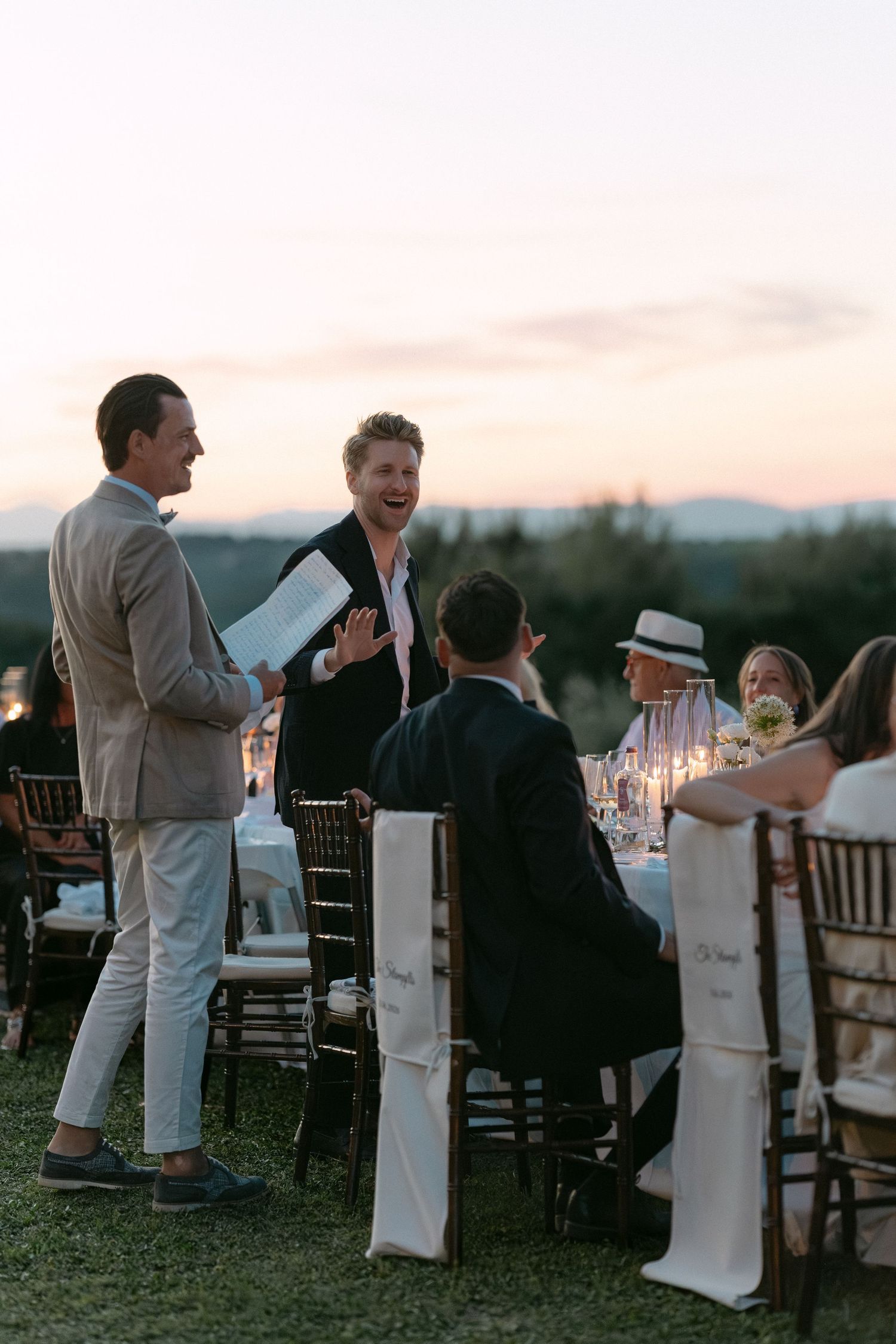 Guests laughing and socializing at an elegant outdoor wedding reception dinner at dusk with candles.