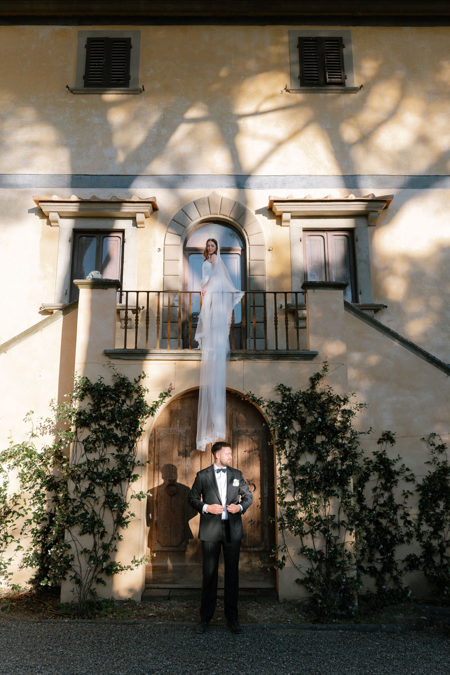 Bride on balcony with flowing veil, groom standing below at elegant Italian villa entrance, romantic wedding photo.