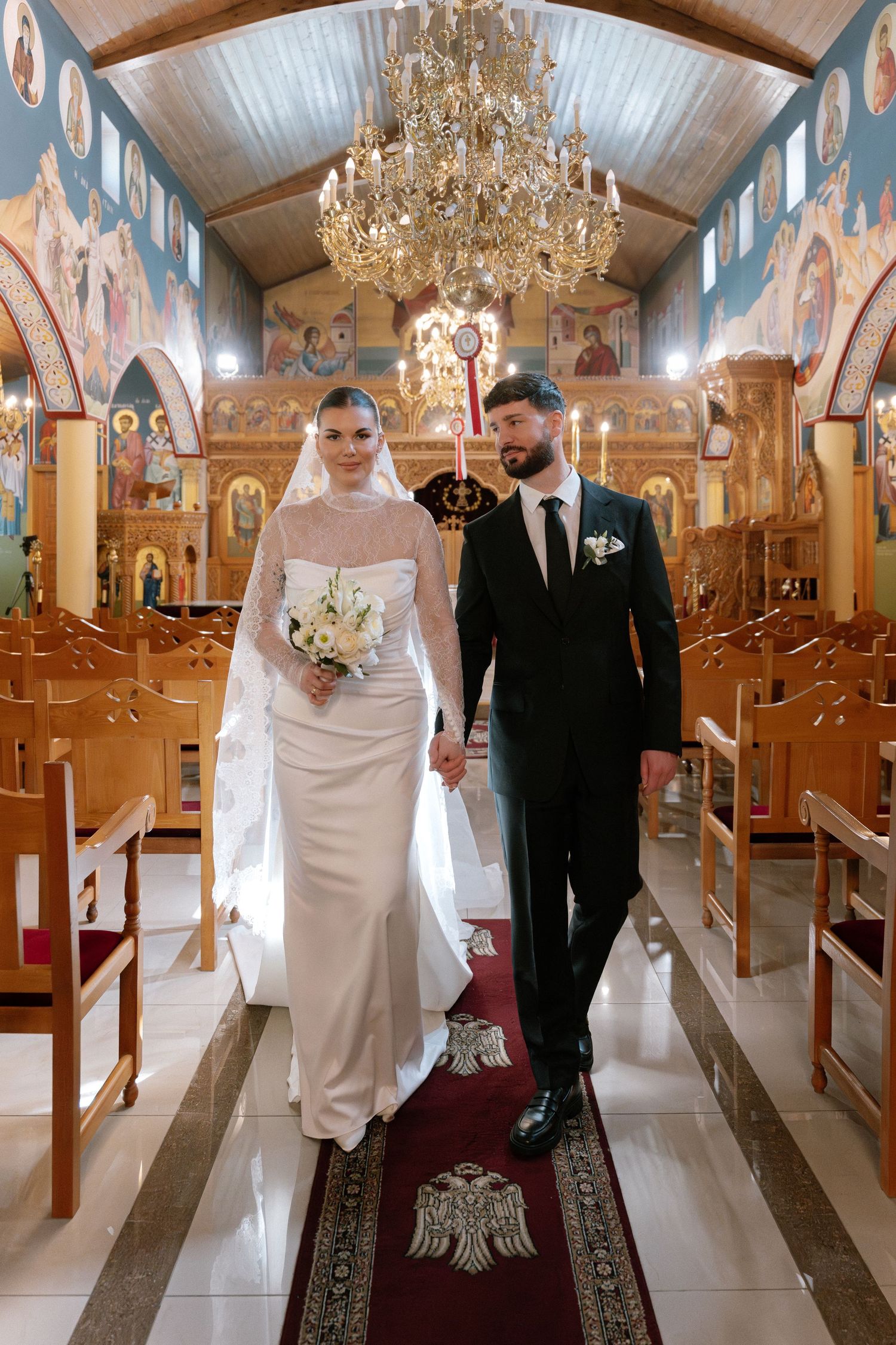 Bride and groom walk down the aisle of an Orthodox church, holding hands, with chandelier and icons above.