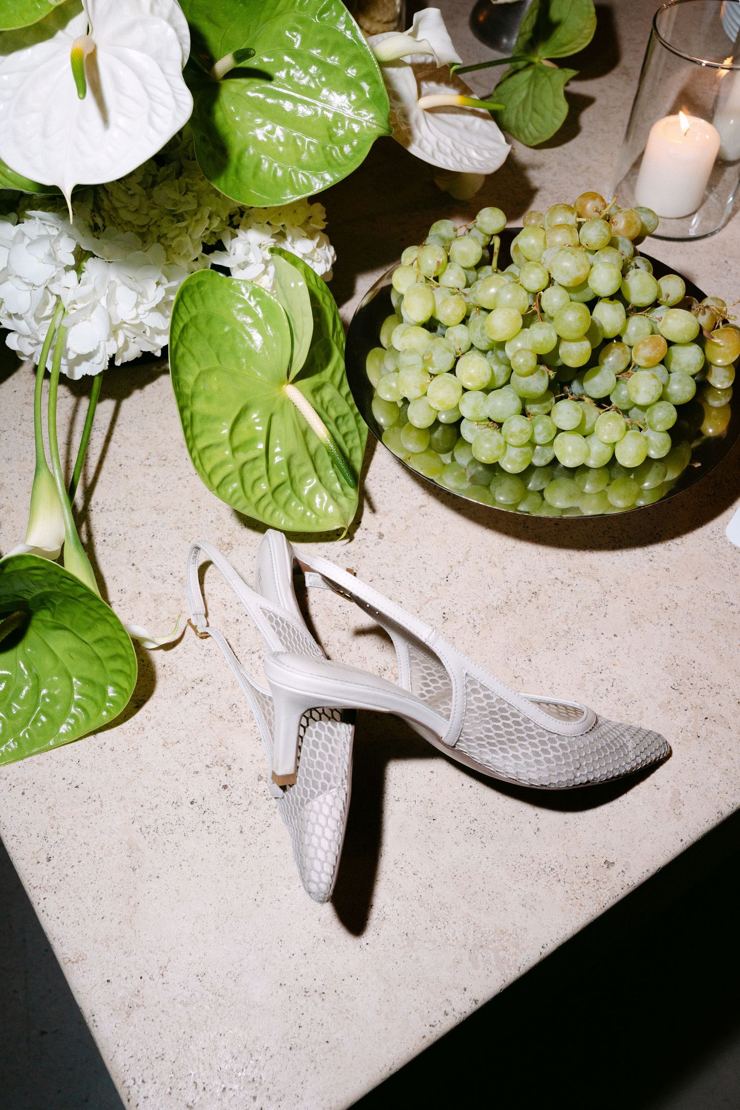 White strappy heels on stone surface with green grapes, anthurium leaves, hydrangeas, and a candle in soft light.