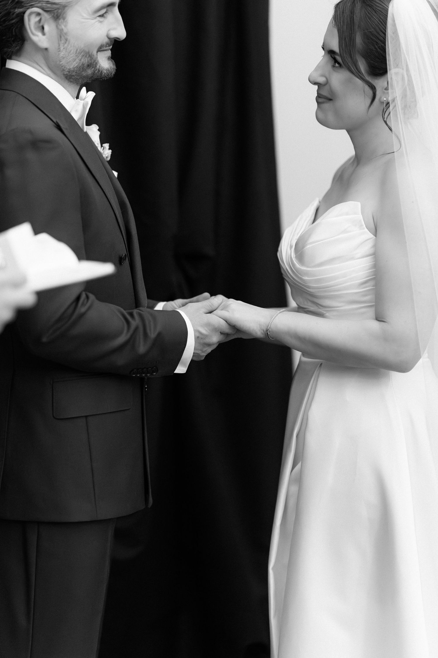 Black and white photo of bride and groom holding hands during wedding ceremony, bride in strapless gown with veil.