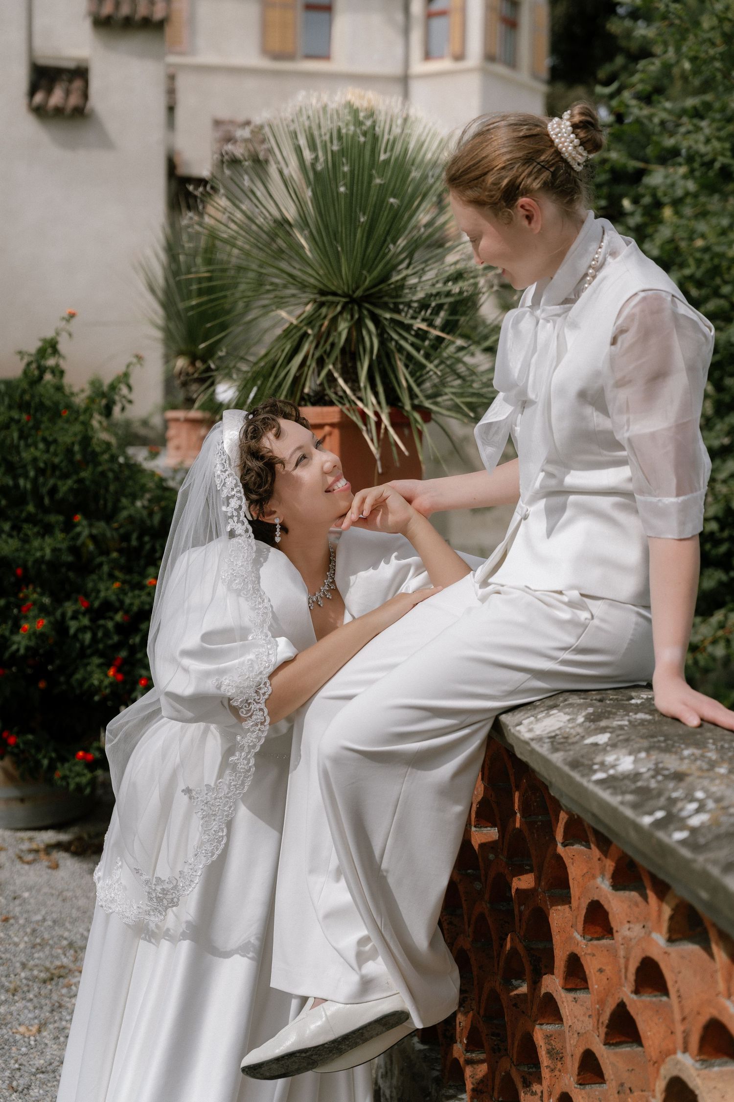 Two brides in white wedding attire hold hands outdoors near lush greenery and a rustic brick wall.