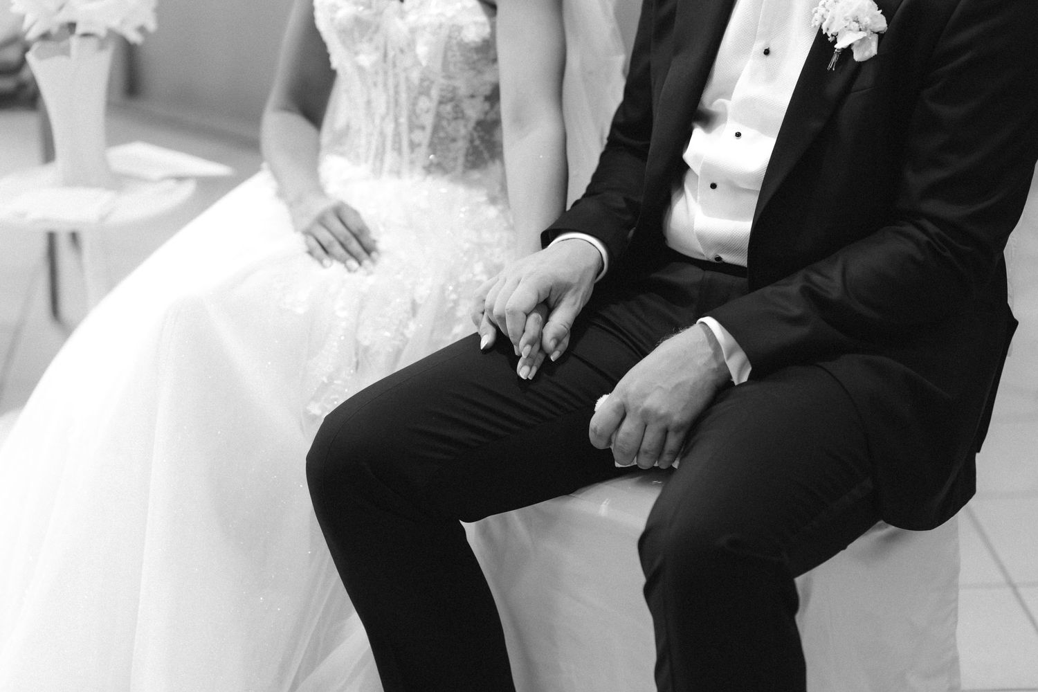 Black and white photo of a seated bride in a lace wedding gown and groom in a suit with boutonniere holding hands during the ceremony.