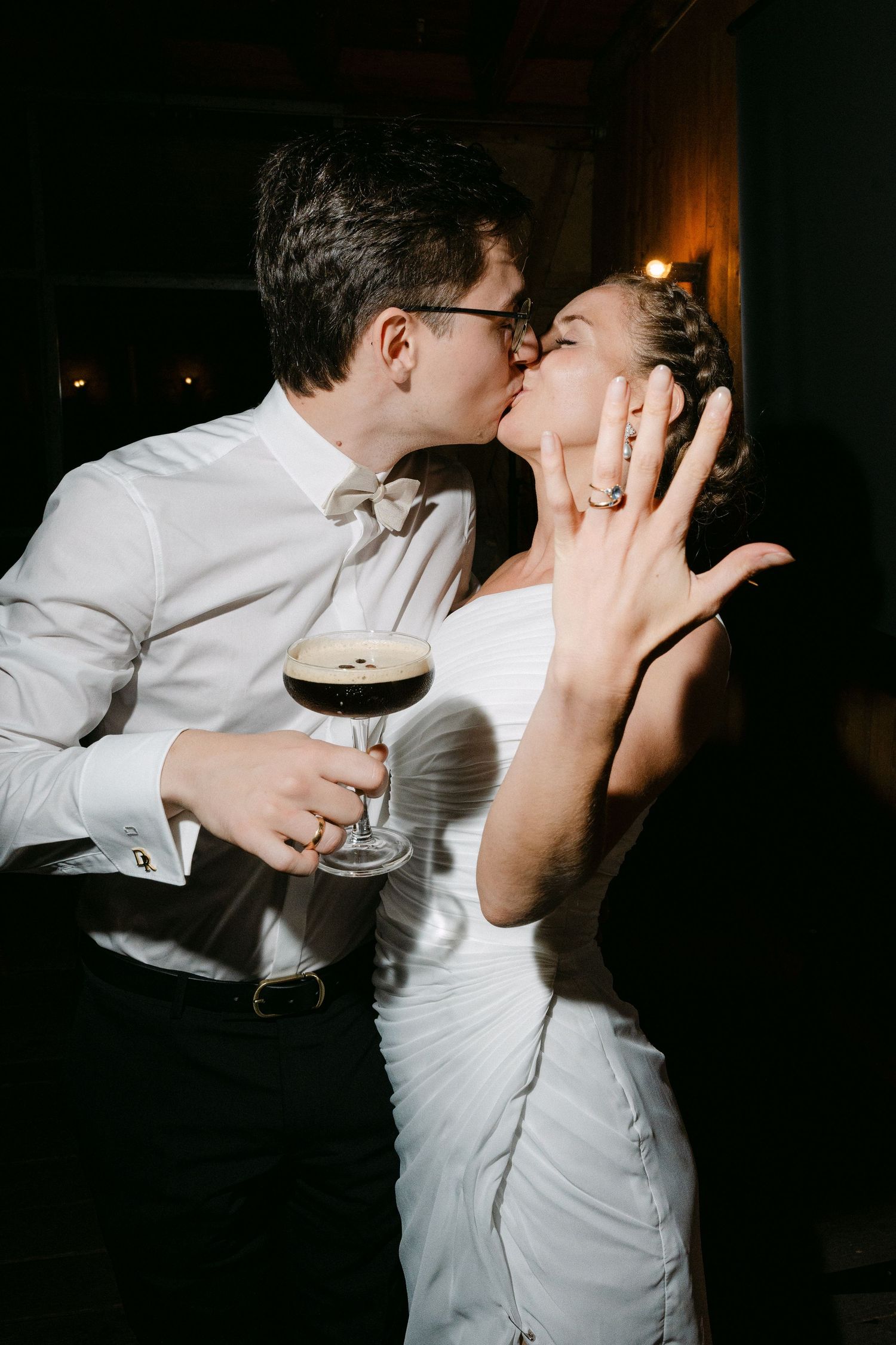 A couple in formal attire kisses while the woman shows off her ring and the man holds an espresso martini in a dimly lit room.