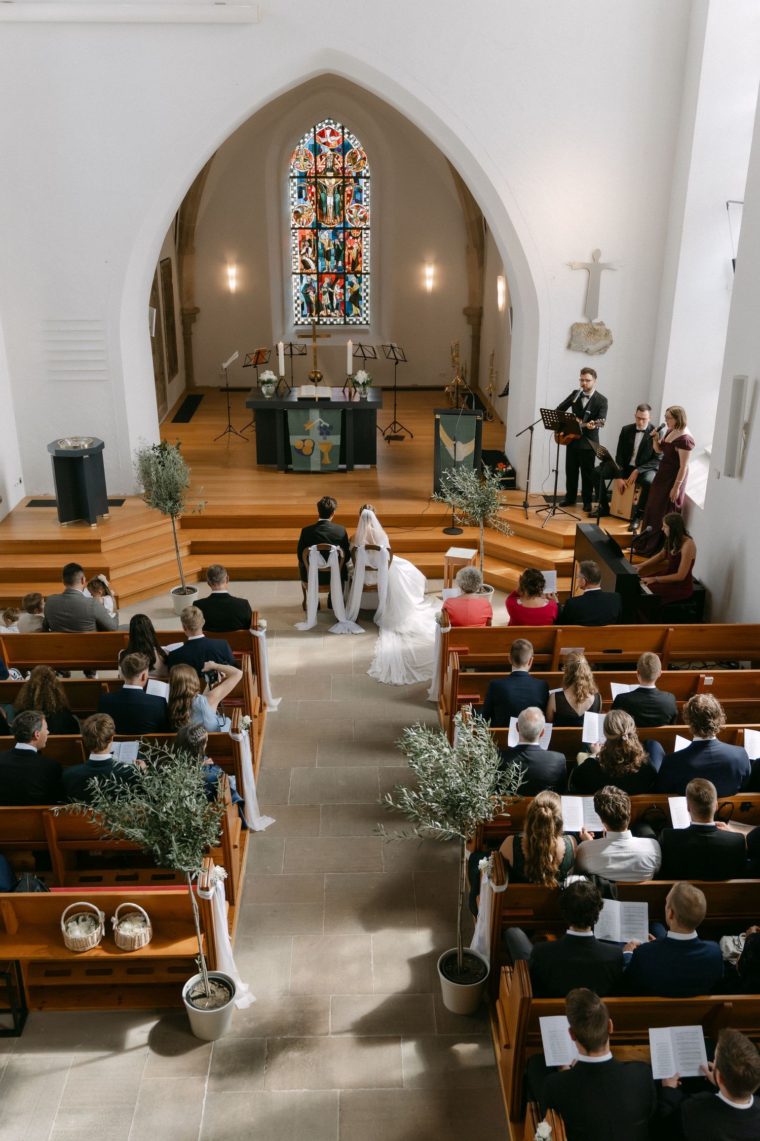 Wedding guests fill the wooden pews of a church as a bride and groom stand at the altar beneath a stained glass window while musicians play.