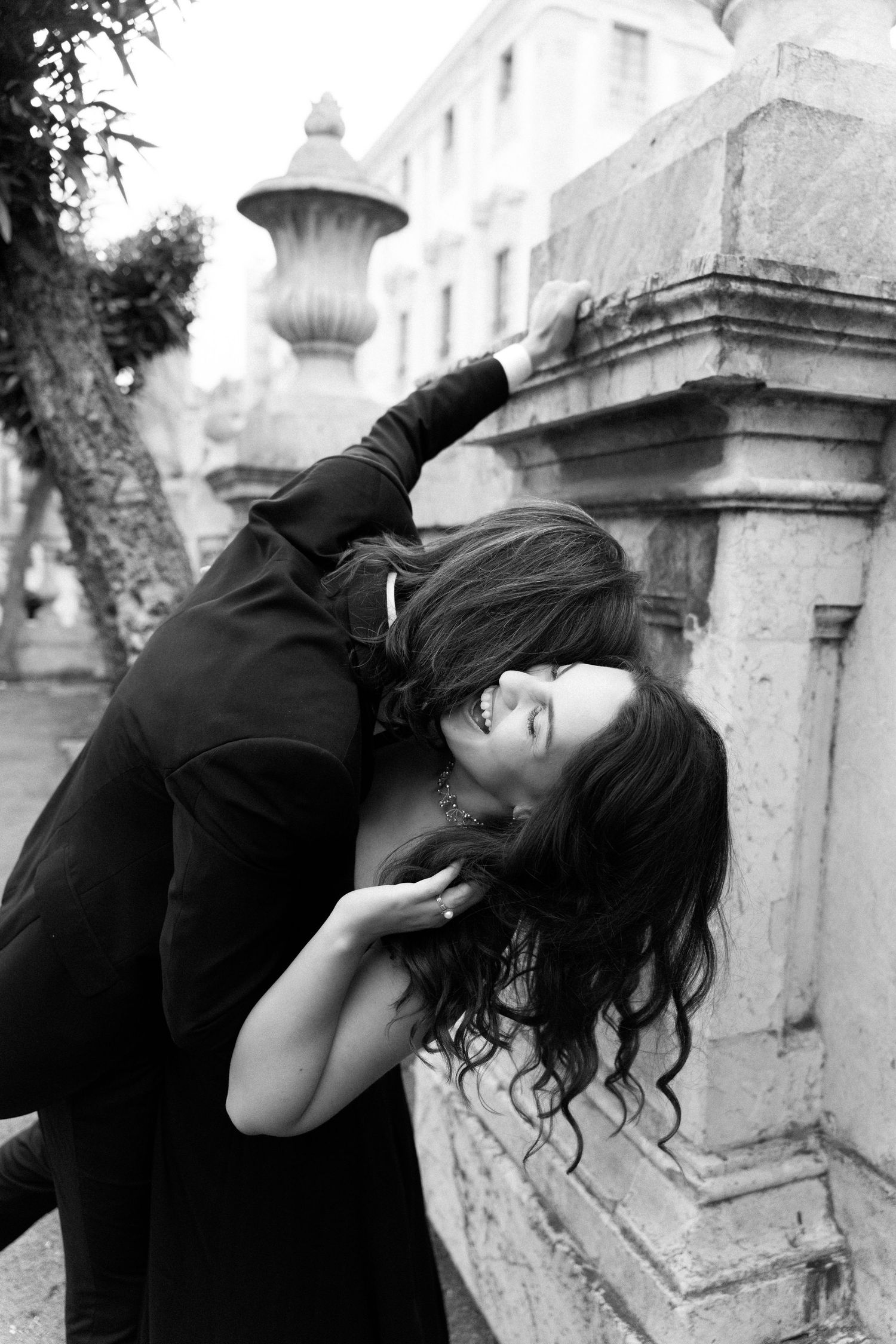 Black and white photo of a romantic couple embracing and kissing near ornate stone pillars in an elegant outdoor setting.