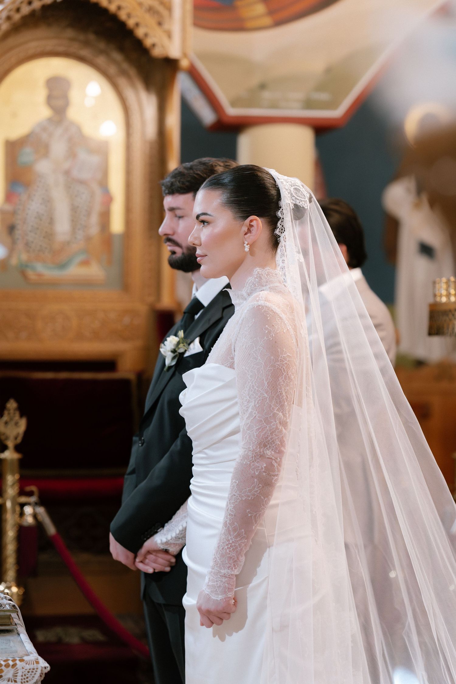 Bride in white lace gown and veil stands beside groom in black suit during Orthodox church wedding ceremony.