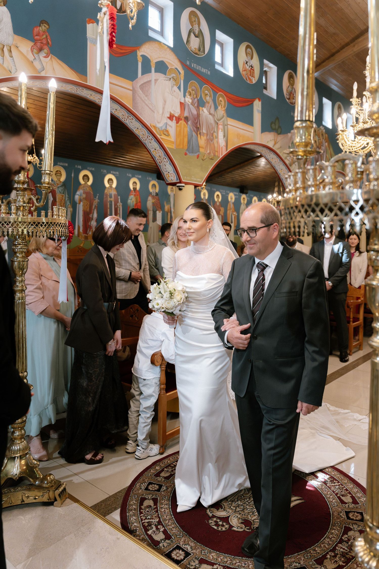 Bride in white gown walks down the aisle with escort in Orthodox church, colorful Byzantine murals in background.