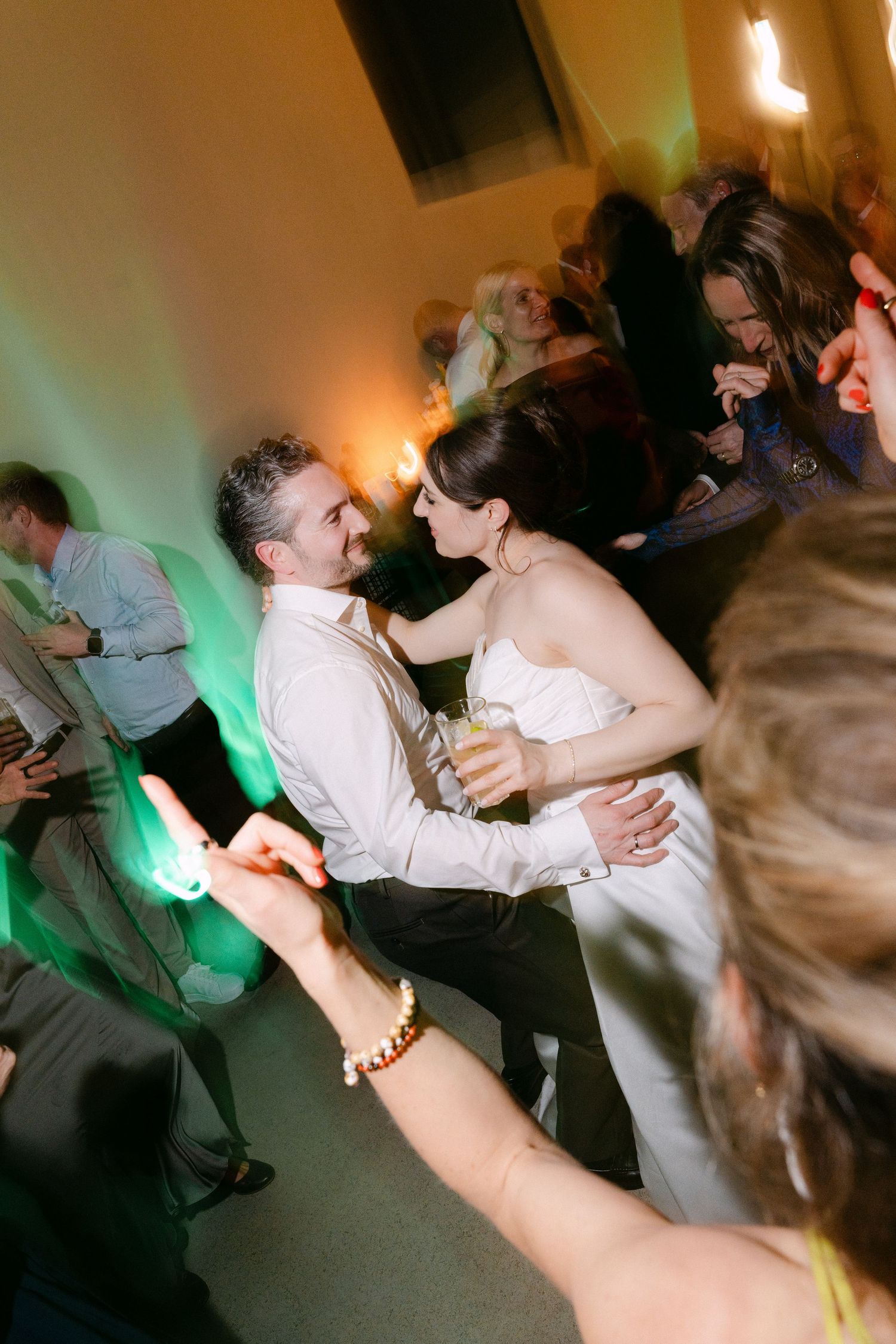 A bride and groom share a romantic dance at their wedding reception, surrounded by guests under warm green ambient lighting.