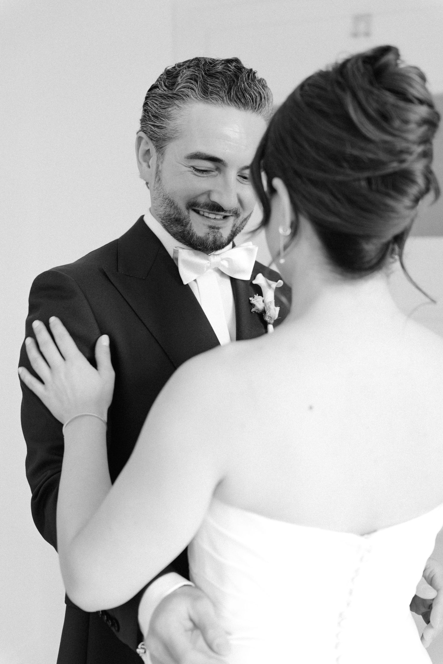 Black and white photo of a smiling groom in a tuxedo embracing his bride during their wedding first look.