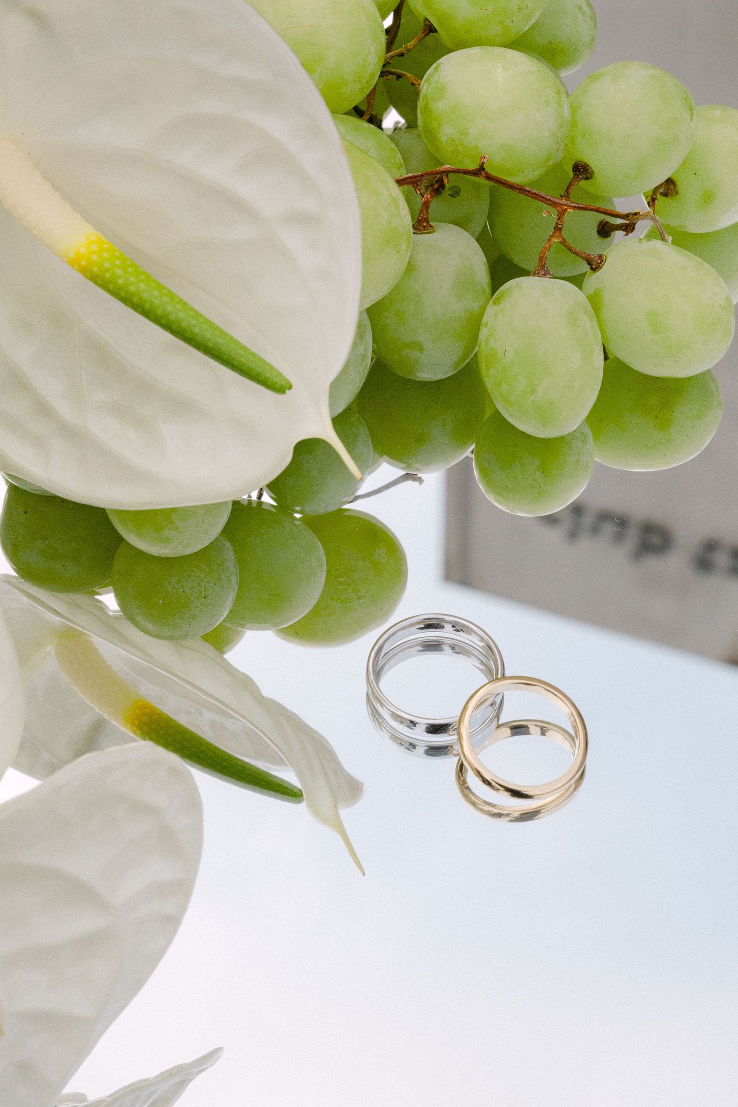 Two wedding rings rest beside white anthurium flowers and fresh green grapes on a white surface.
