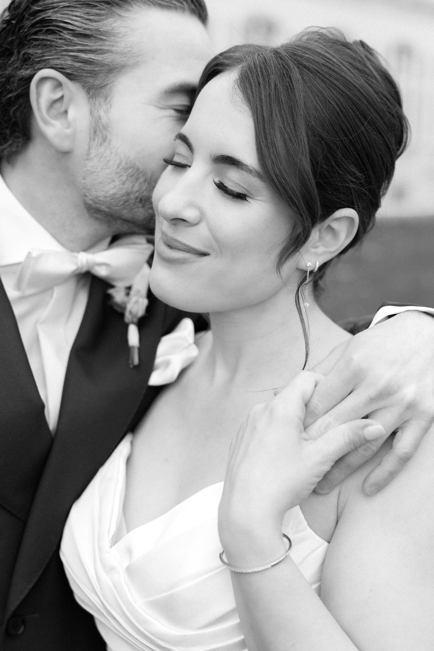 Black and white wedding photo of groom kissing bride on cheek, bride smiling with eyes closed, wearing white gown.
