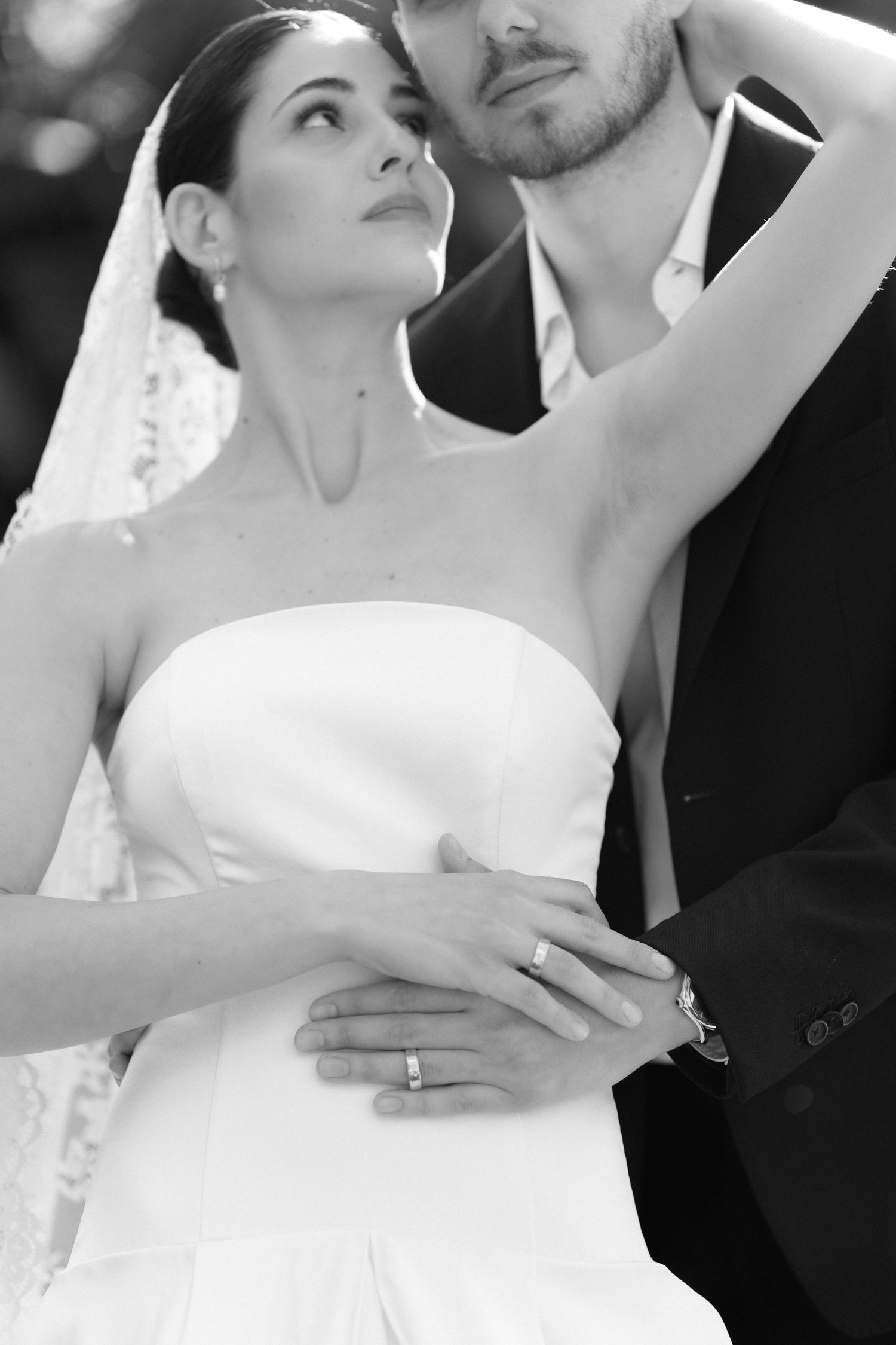 Black and white photo of bride in strapless gown and groom in suit embracing, showing wedding ring on her hand.