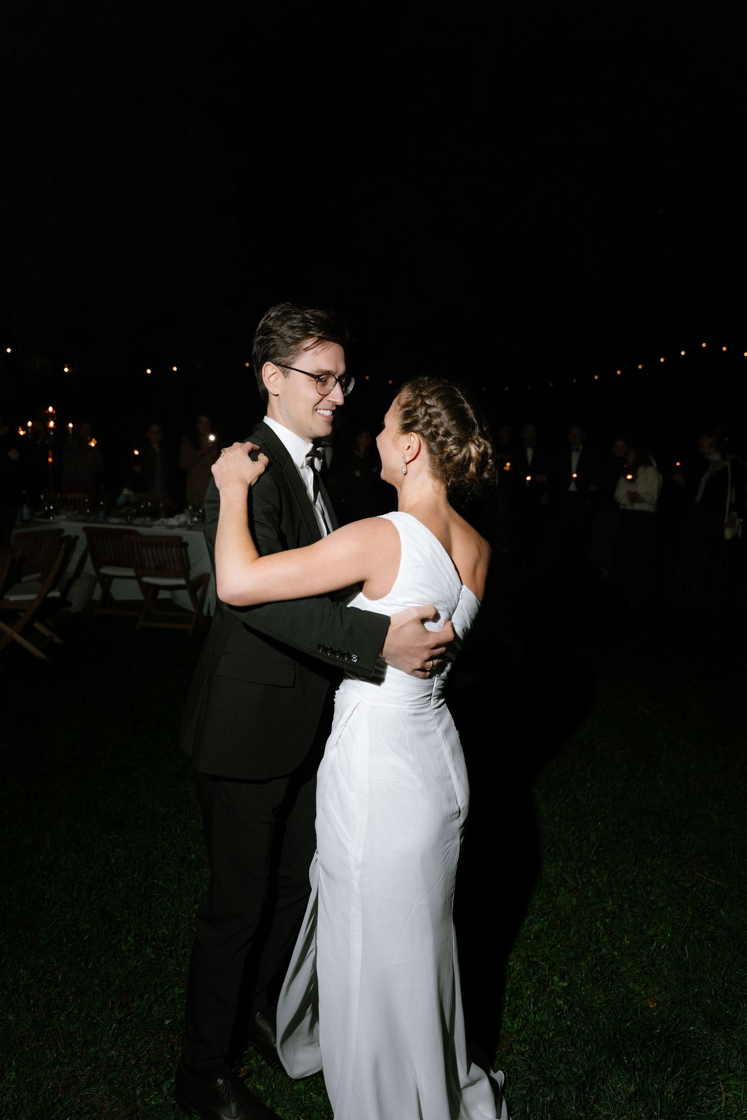 Bride in a white gown and groom in a black suit and glasses share a slow dance outdoors at night during a wedding reception.