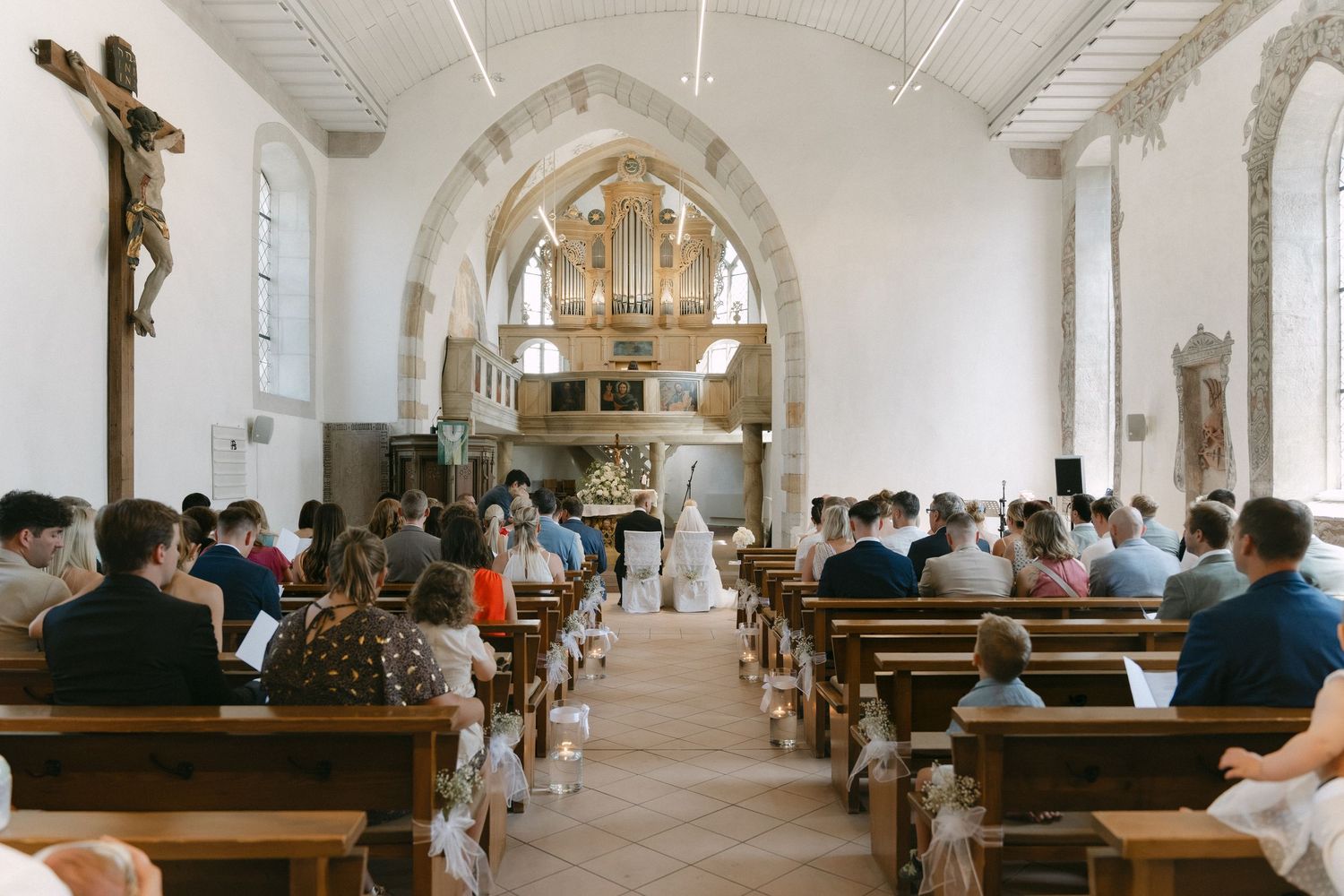 Wedding guests seated in wooden pews watch a bride and groom at the altar of a bright historic church with arched ceilings and a crucifix.
