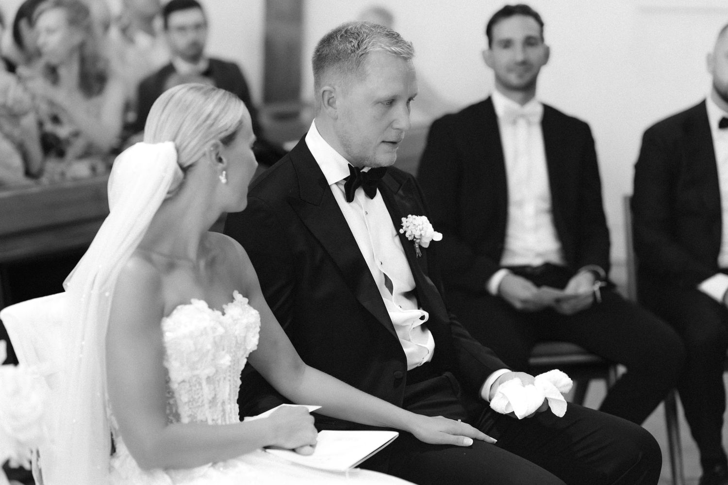 Black and white photo of a bride in a strapless lace gown and a groom in a tuxedo with bow tie seated together during a wedding ceremony.
