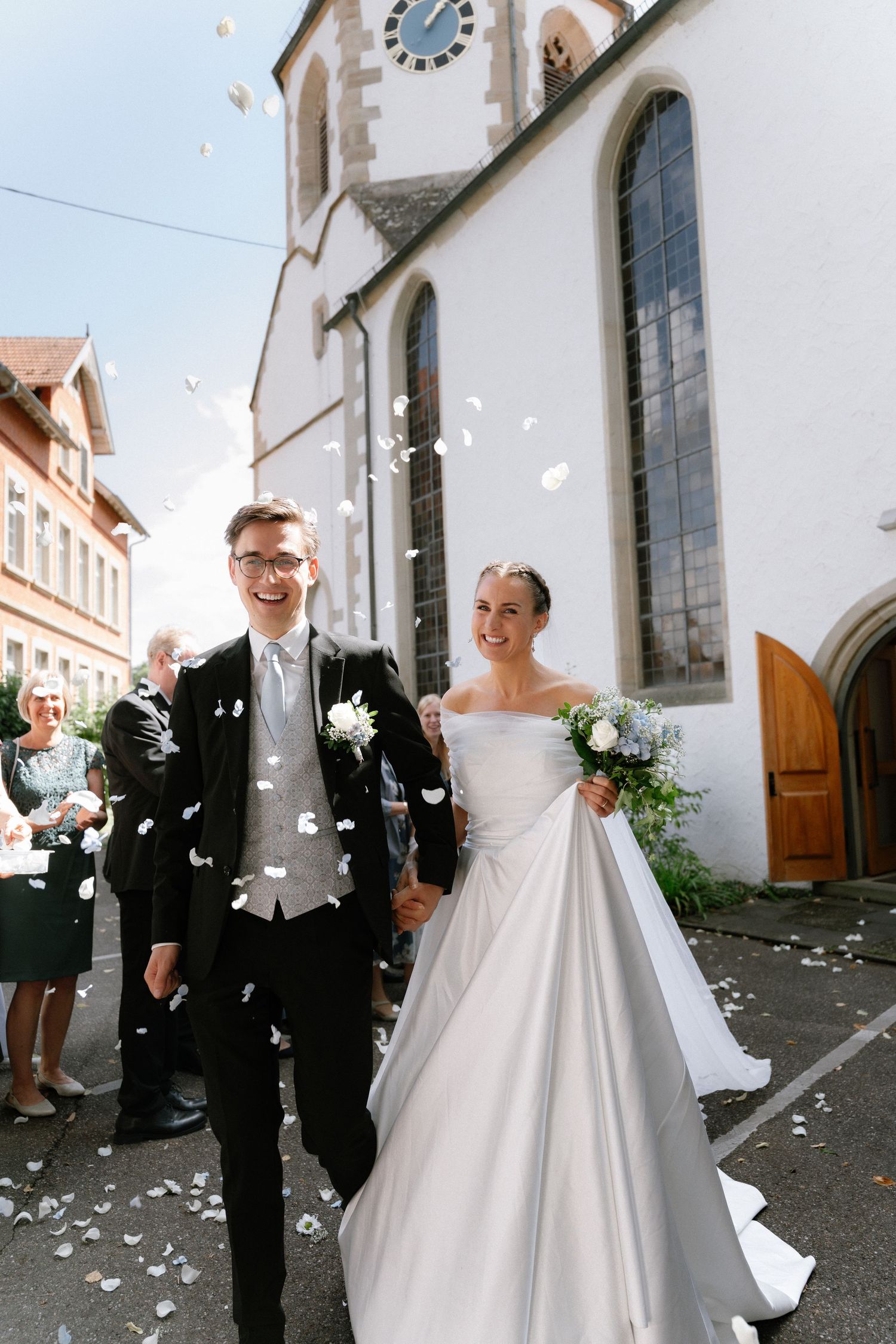 Smiling bride in a strapless satin gown and groom in a black suit walk hand in hand outside a church as guests toss confetti.